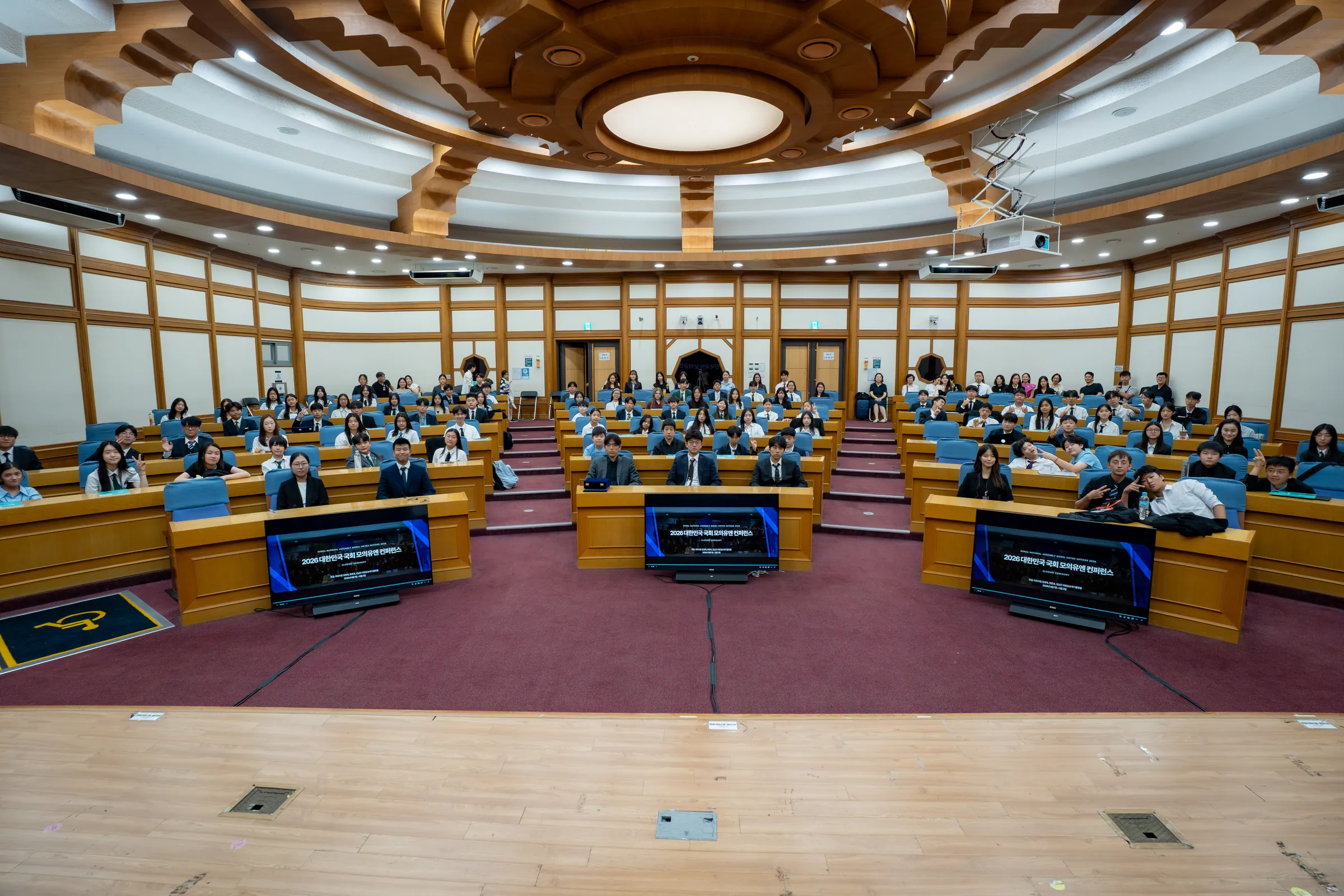 Wide view of a tiered, wood-panelled assembly hall with an ornate ceiling, filled with seated student delegates at curved desks.