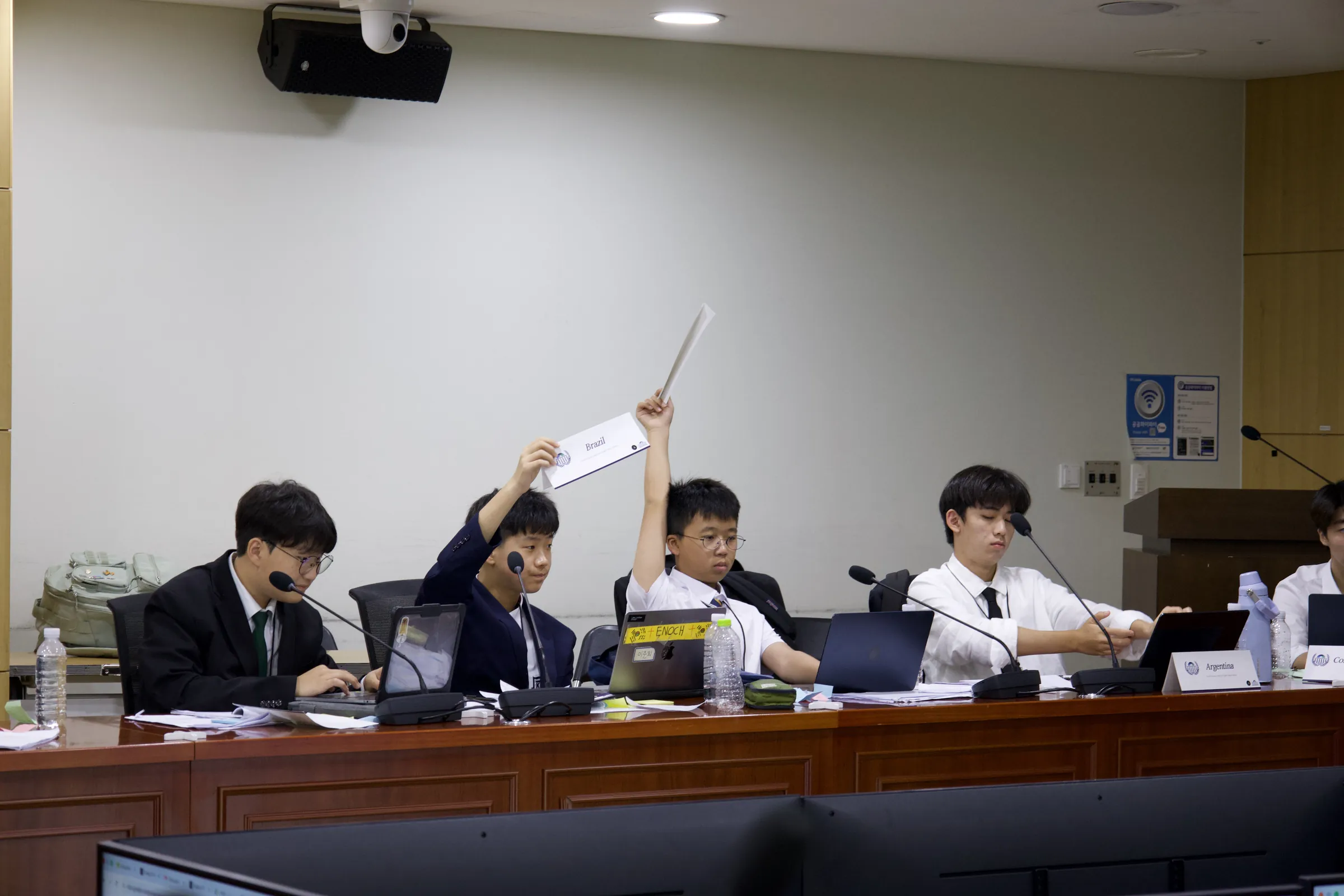 Delegates in suits seated at a curved committee table with microphones and laptops; two of them raise their country placards high.