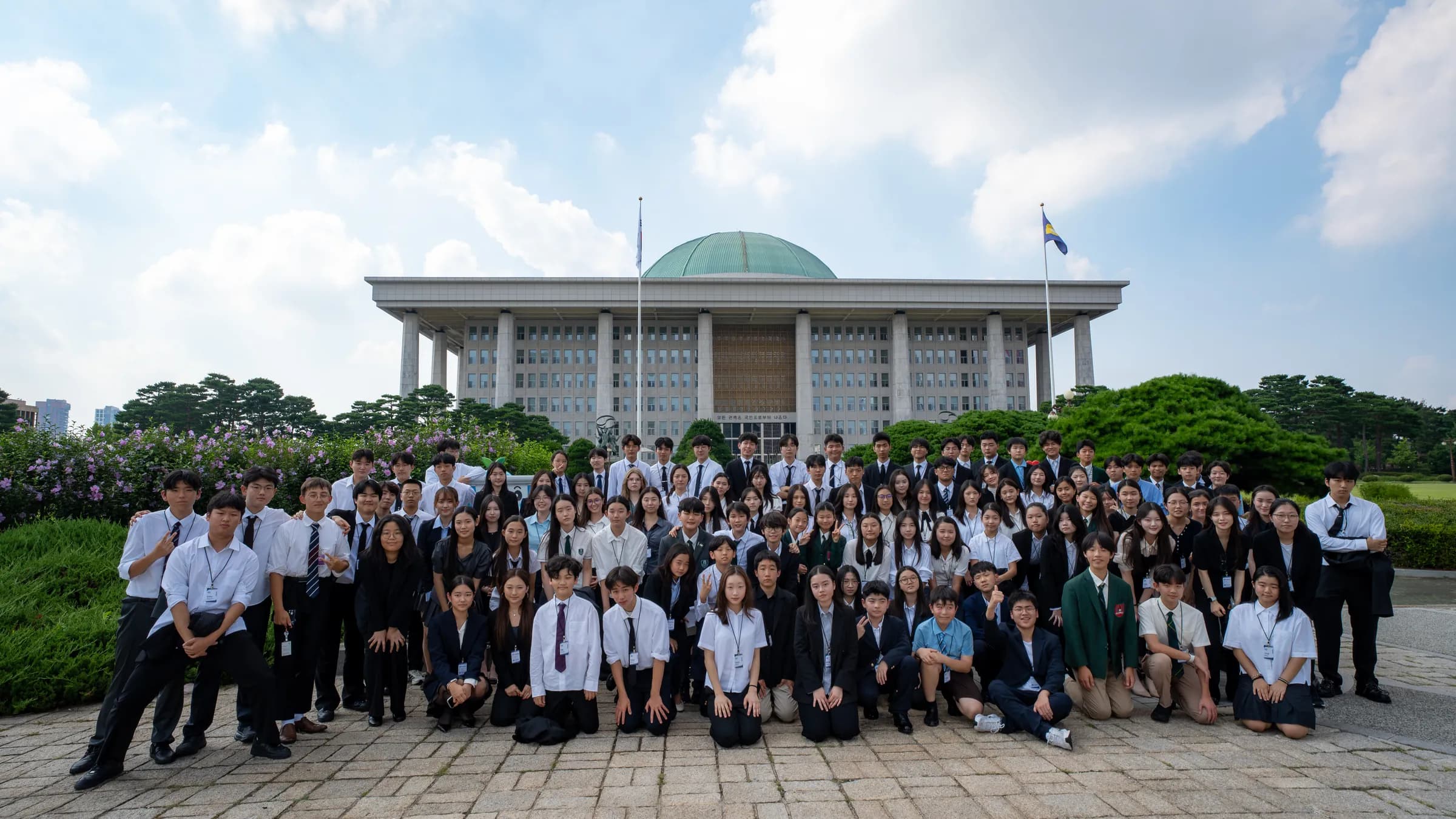 A large group of students in suits poses on the lawn in front of the domed National Assembly building in Seoul.