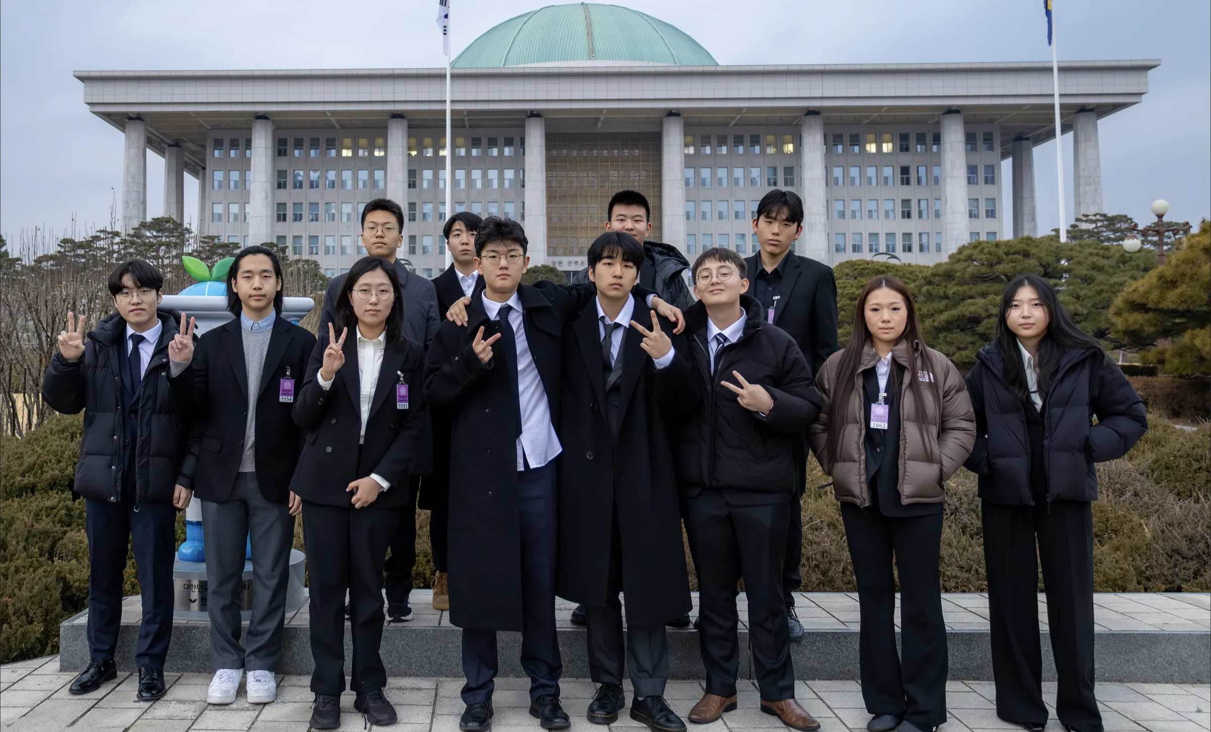 A group of students in dark coats and suits poses outdoors in front of the domed National Assembly building.