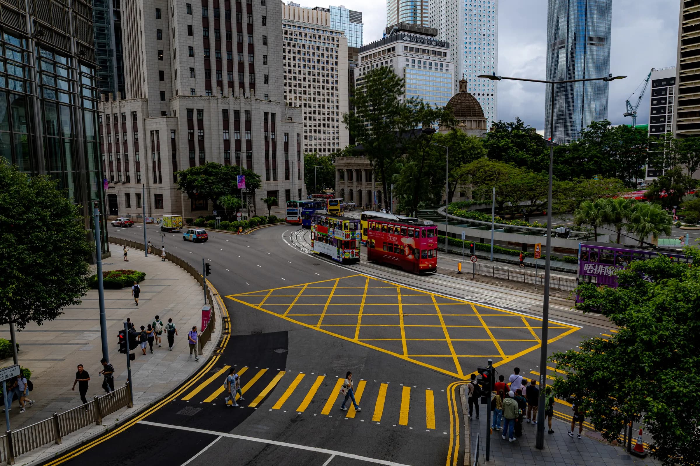 Central from a footbridge: a yellow box junction, red double-decker trams, a domed colonial building in the trees and glass towers behind.