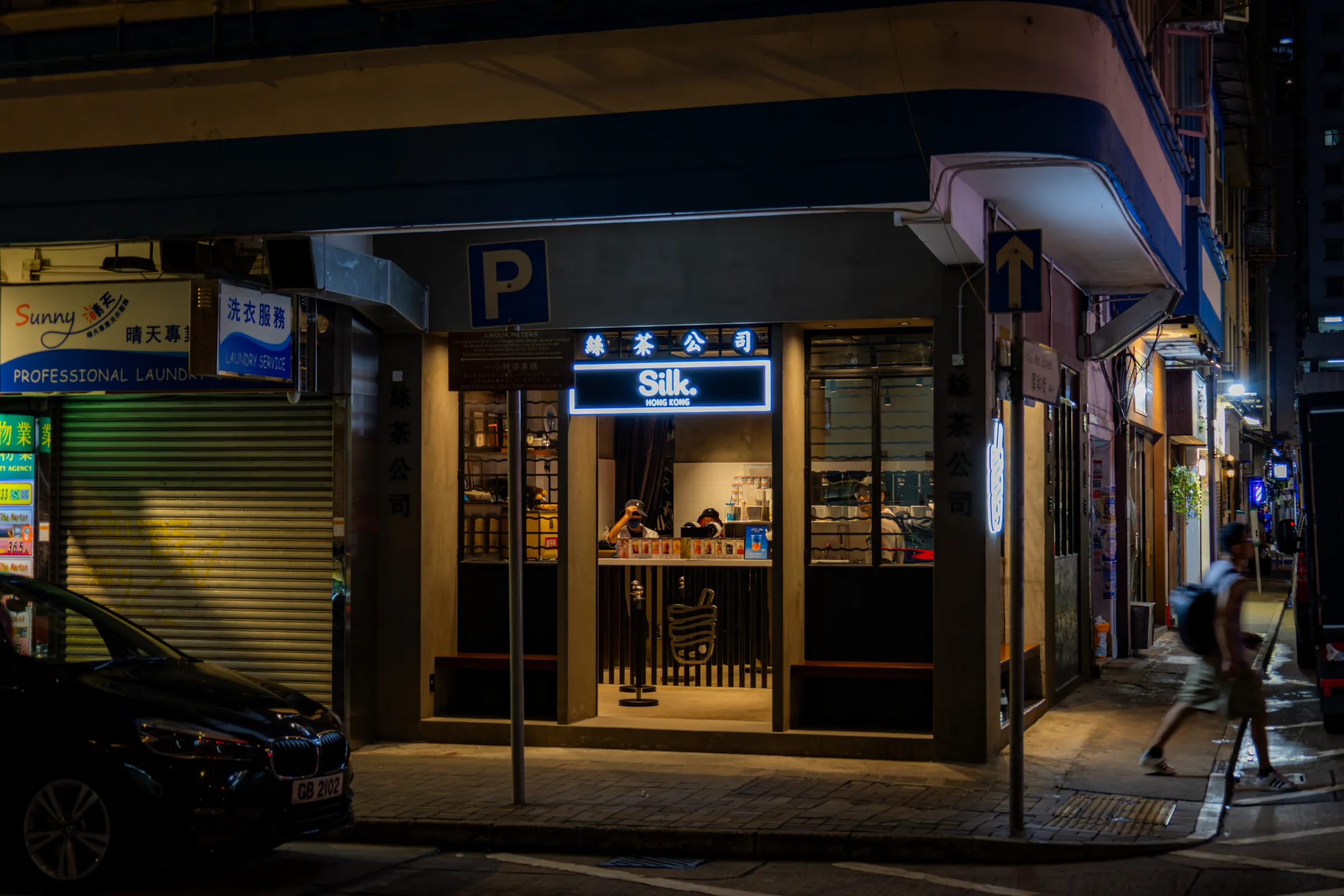A corner shopfront in Hong Kong at night, warm light inside under a sign reading Silk, a parked car at left and a figure walking past on the pavement.