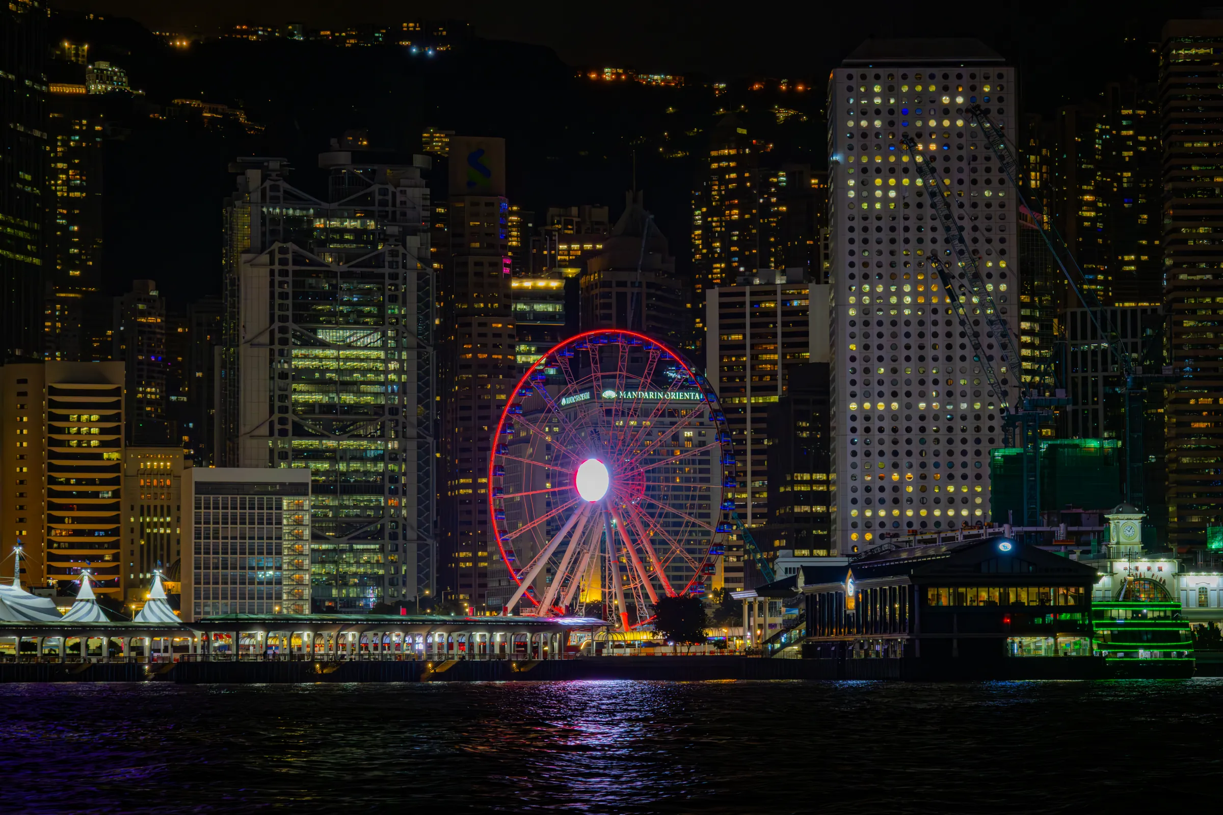 The Hong Kong Observation Wheel ringed in red light on the Central waterfront at night, office towers dark behind it and the harbour in front.