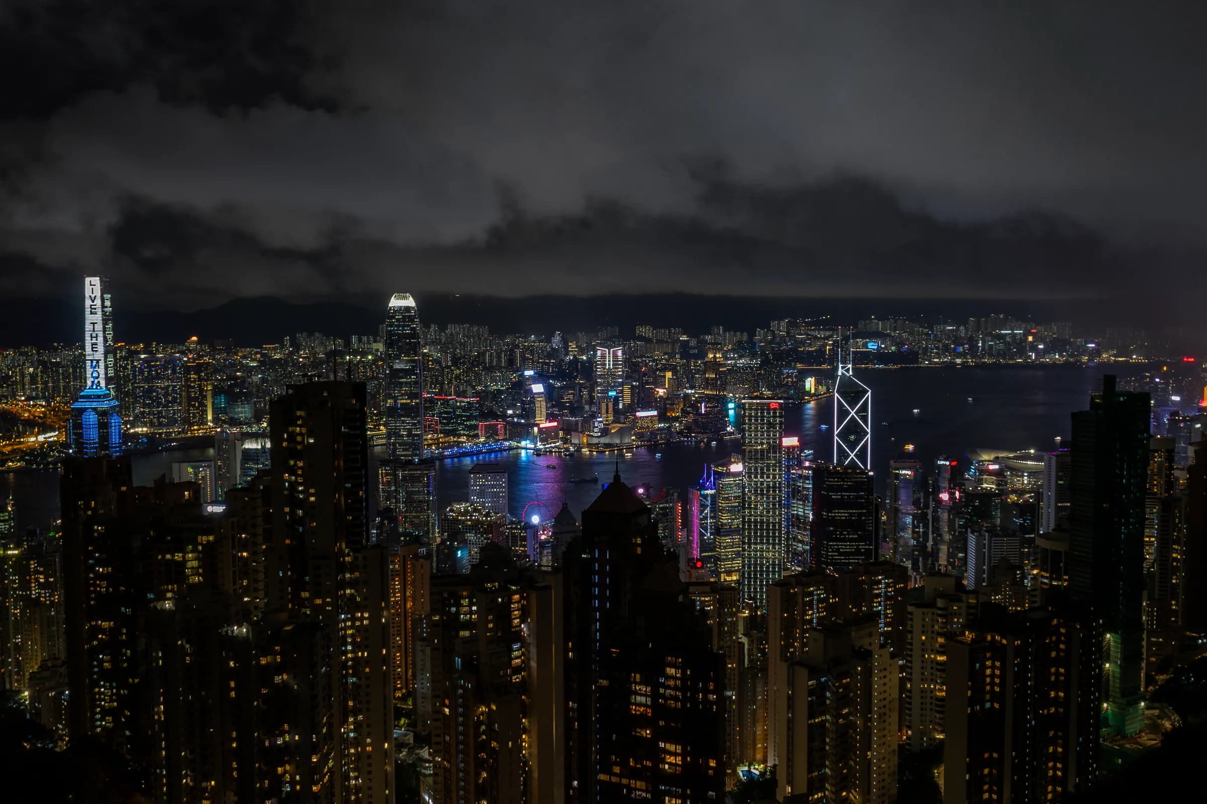 Victoria Harbour from the Peak at night: the ICC and IFC towers lit above the dense lights of both shores, heavy cloud over Kowloon.