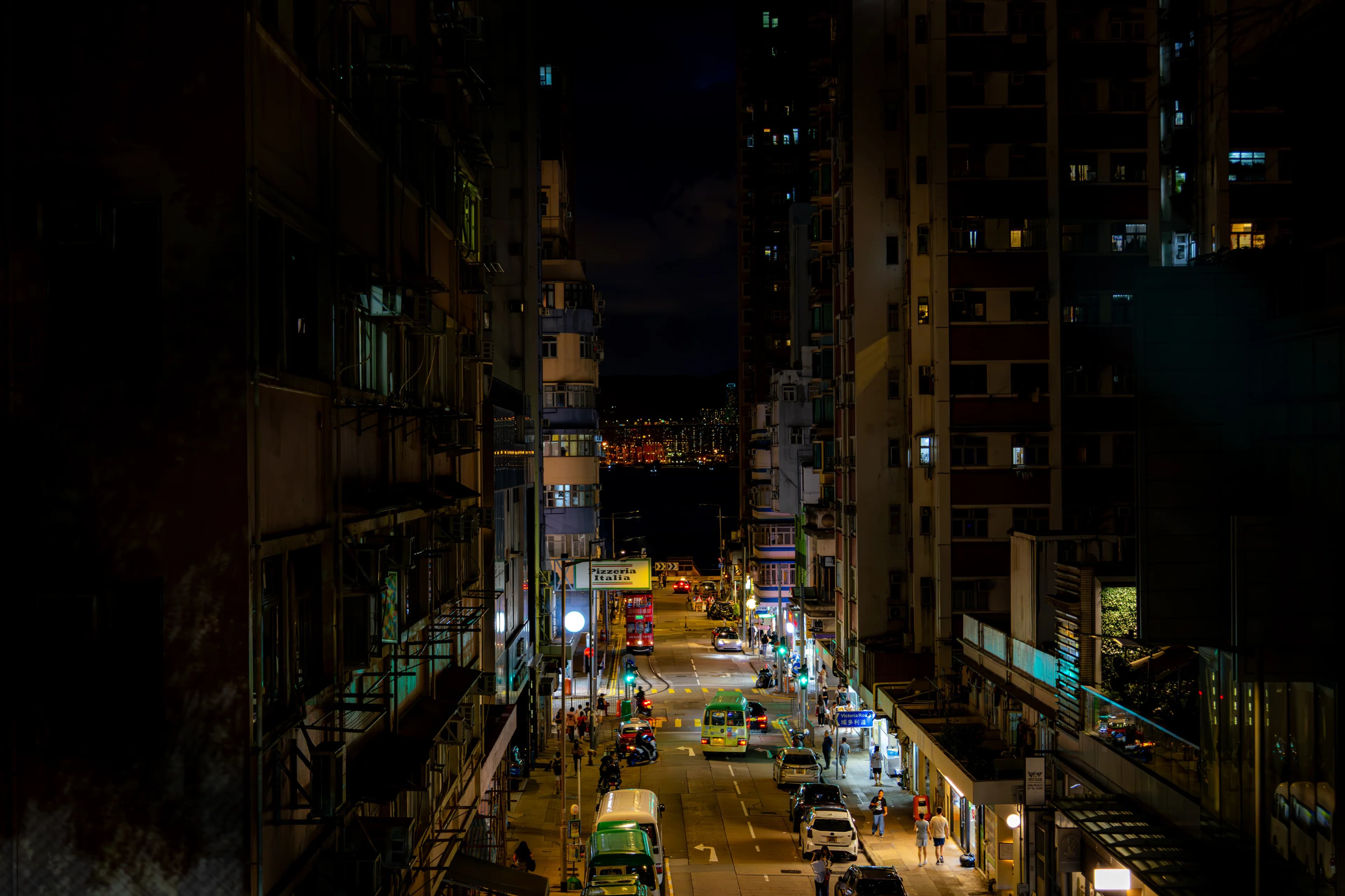 A narrow street at night from high up between apartment blocks, green minibuses and headlights running down to the harbour.