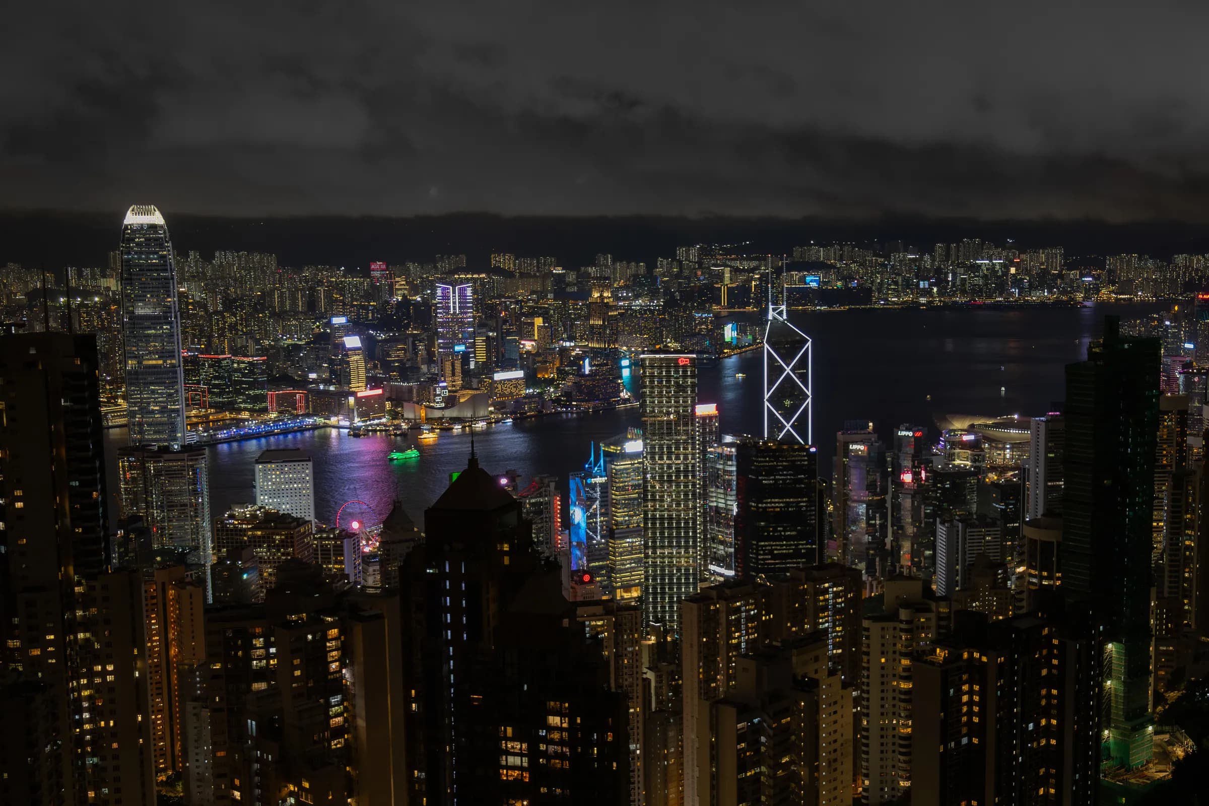 Victoria Harbour from the Peak after dark: towers packed down to the water on both shores, the Bank of China Tower's angles lit in white, and low cloud over Kowloon.