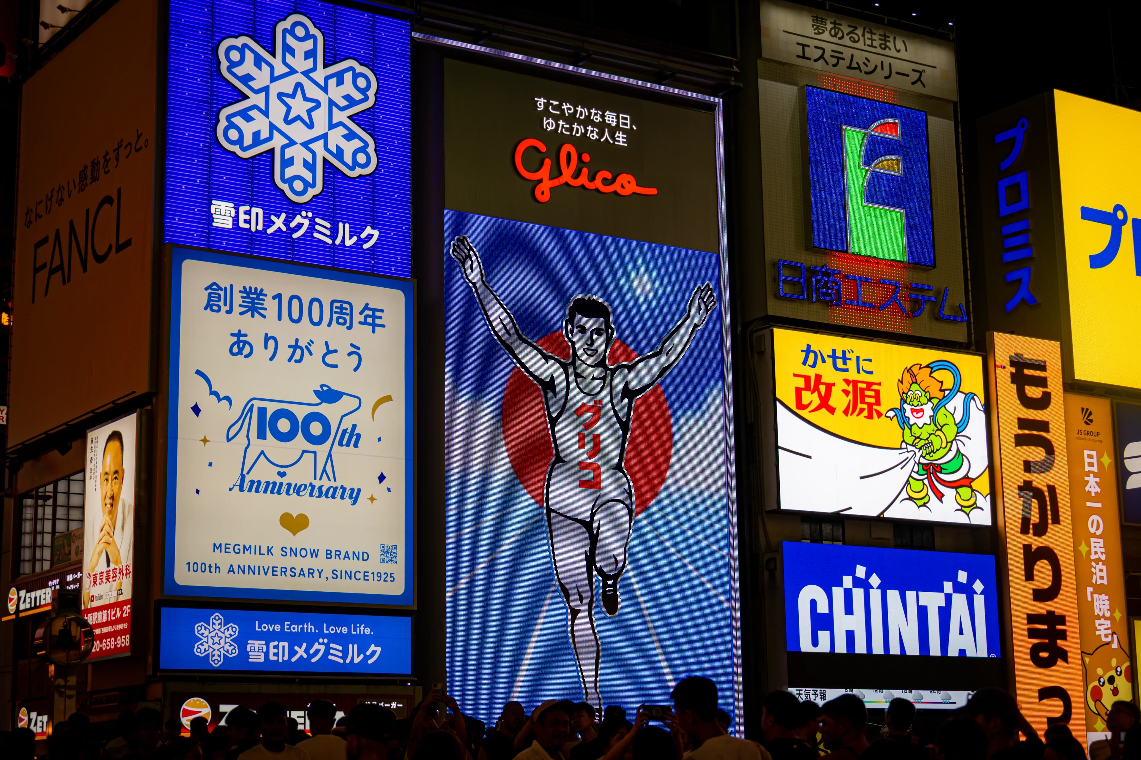 The Glico running man sign in Dotonbori, arms raised against a blue field, surrounded by bright billboards stacked up the buildings.