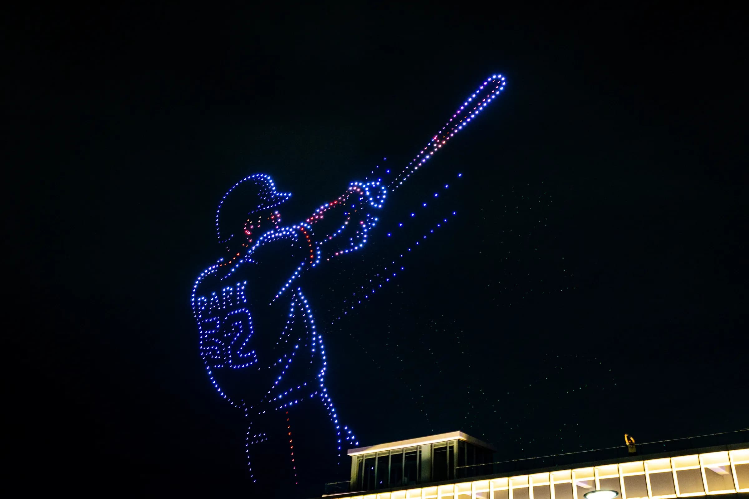 A drone light show over the Han at night: hundreds of blue lights draw a baseball batter mid-swing above the roof of a riverside building.