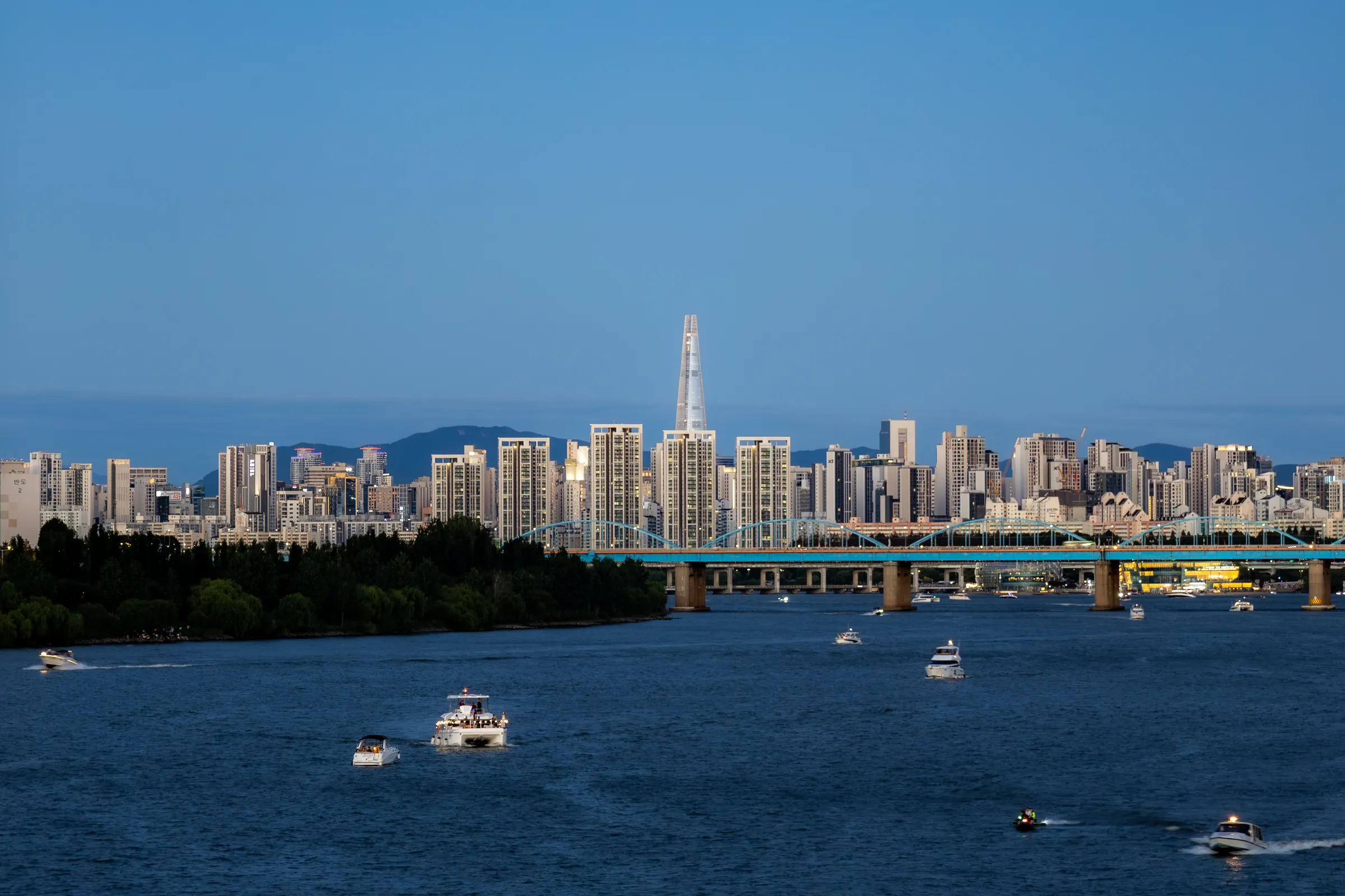 Lotte World Tower rising above the apartment blocks of Jamsil under a clear blue sky, small boats leaving white wakes on the Han in the foreground.