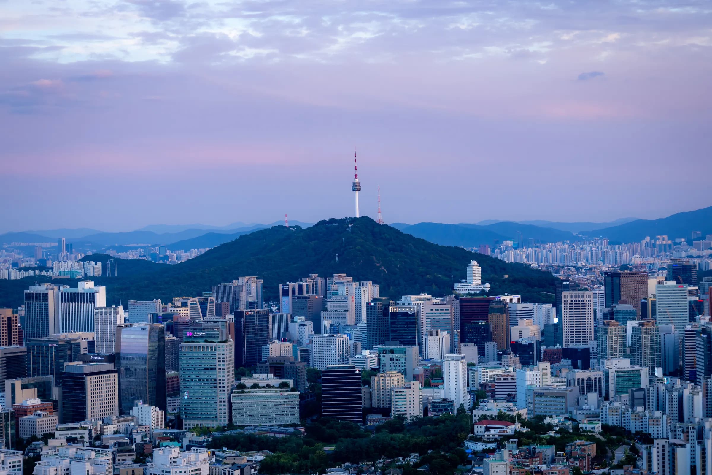 Namsan and N Seoul Tower rising above the dense buildings of downtown Seoul under a soft pink and blue sky, mountains on the horizon.