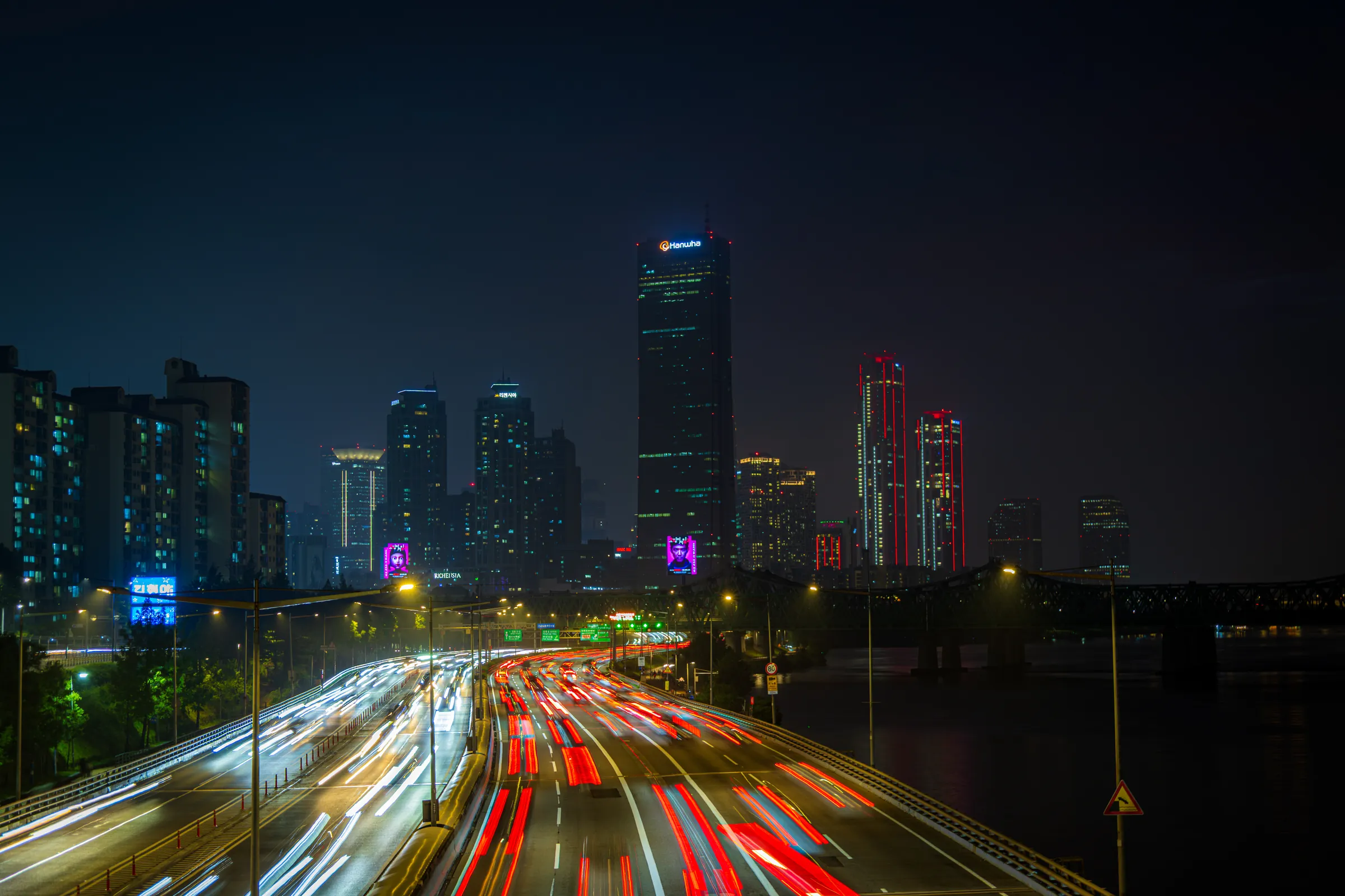 Night over a riverside expressway in Seoul: two seconds of headlights and taillights curve toward a cluster of lit towers, the tallest topped with a glowing sign.