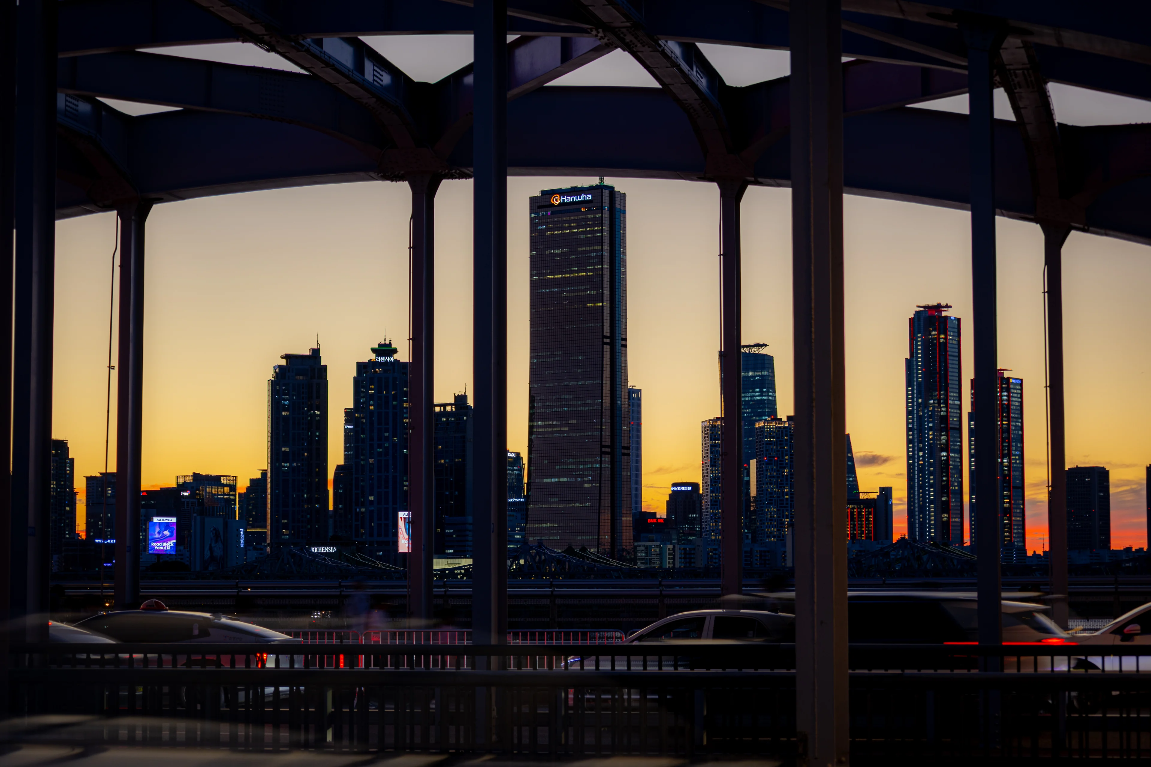 Seoul towers in silhouette against an orange dusk, seen through the dark steel arches of a bridge.