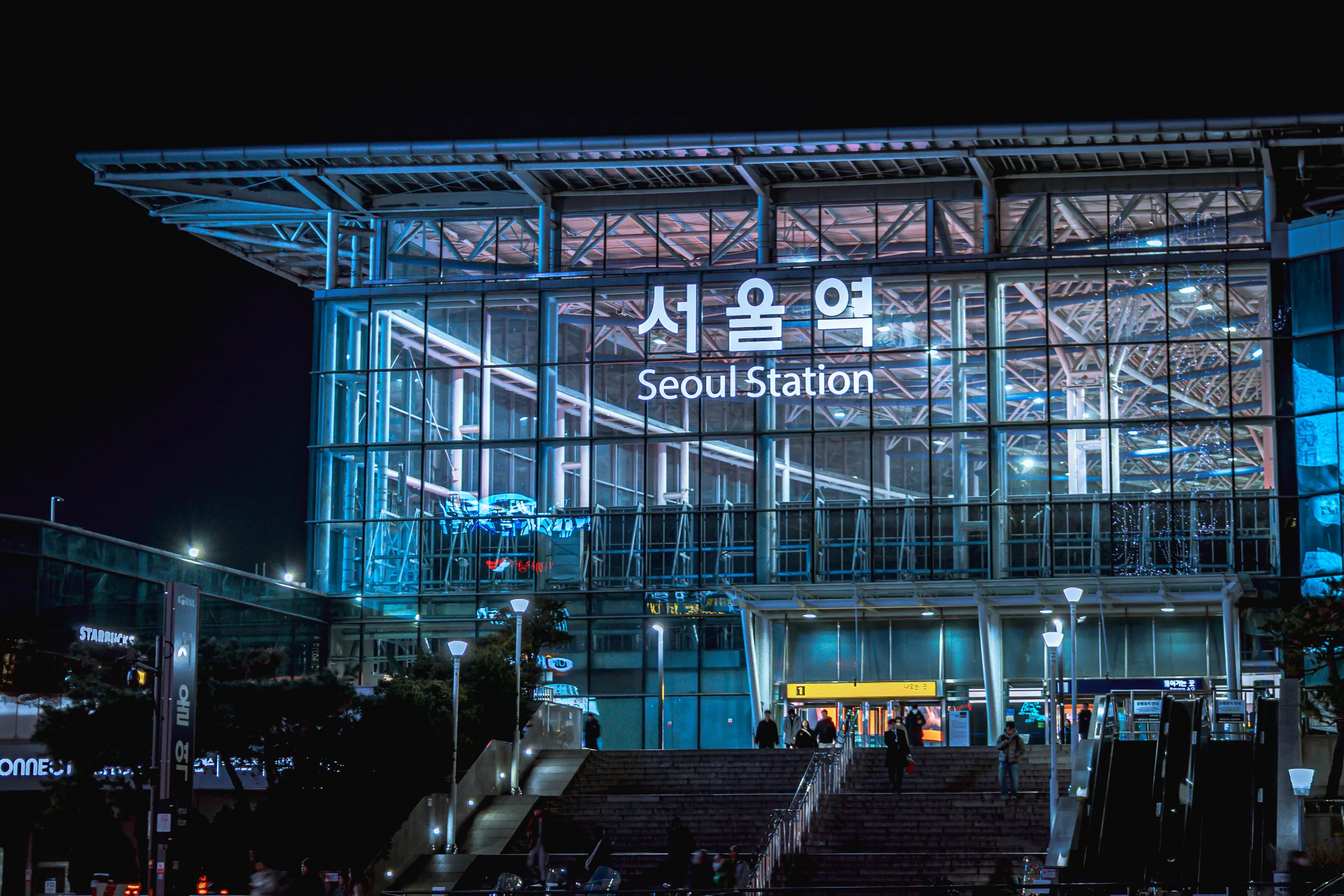 The glass front of Seoul Station at night, lit in cool blue with the station's name in Korean and English across it, a few people on the steps below.