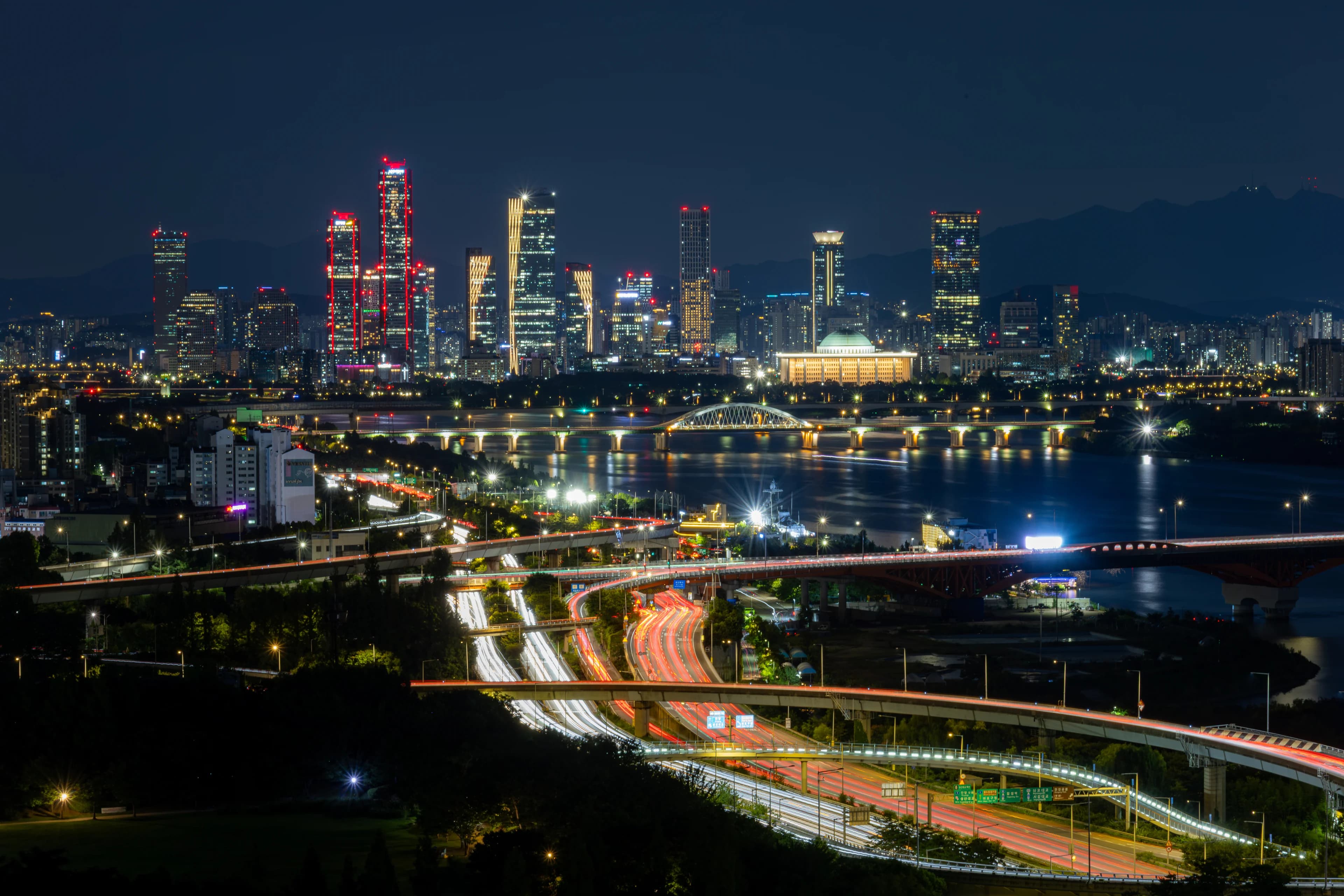 Yeouido at night from a hillside across the Han: towers outlined in red and gold lights, the domed National Assembly building lit below them, a bridge reflected in the river, and expressway ramps in the foreground traced with red and white light trails.