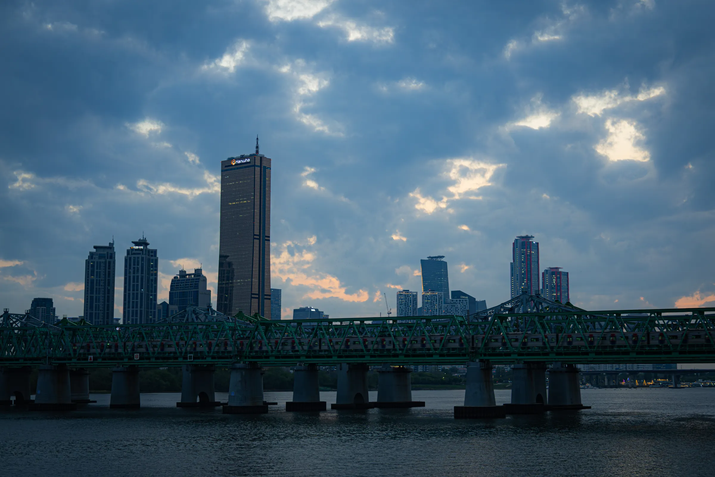 Yeouido's towers in silhouette under a broken evening sky, a green steel railway bridge on stone piers crossing the Han in front of them.