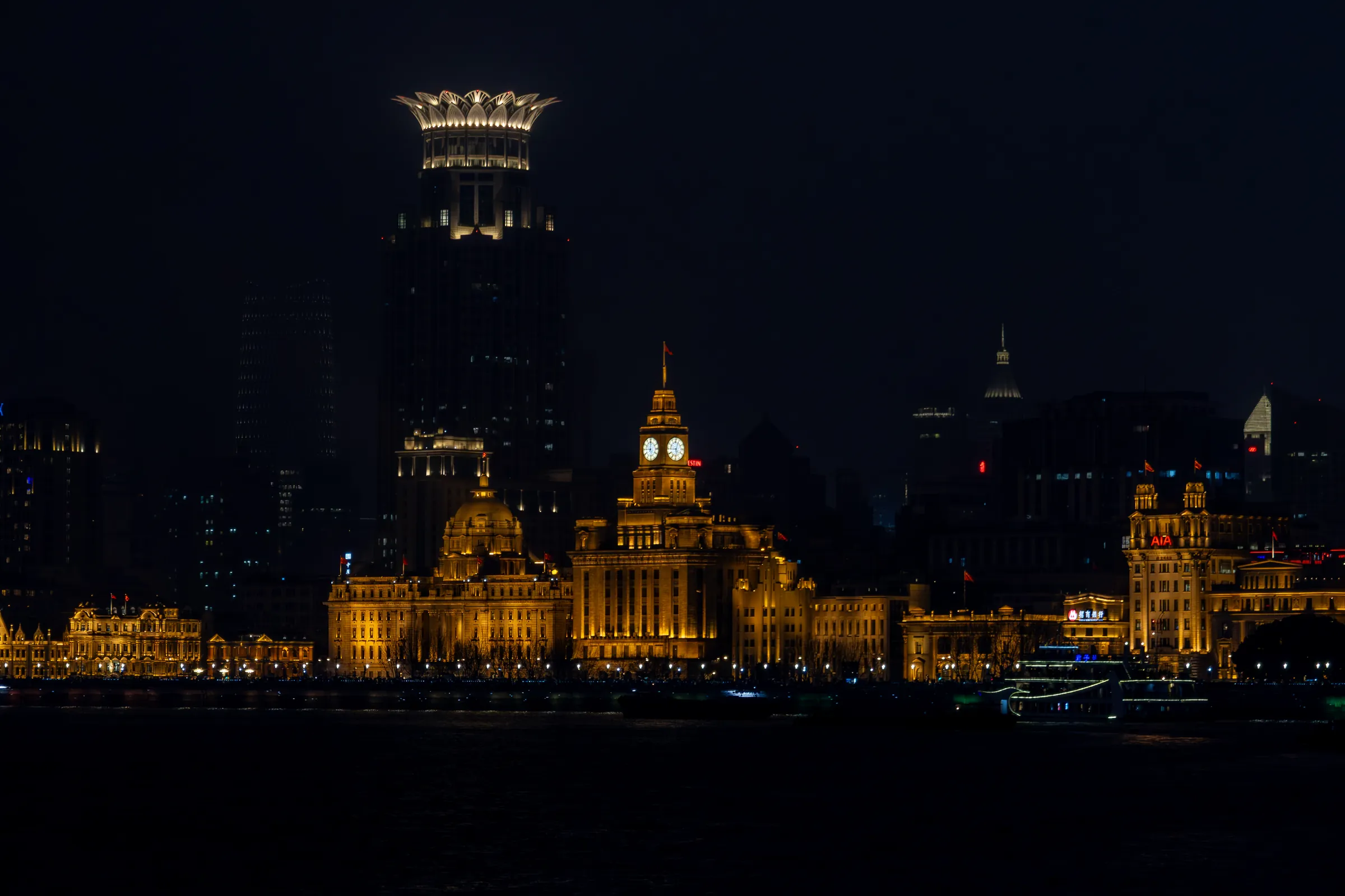 The Bund at night seen across the dark Huangpu River: a domed bank building and the Customs House clock tower glow gold, a crown-topped tower lit behind them.