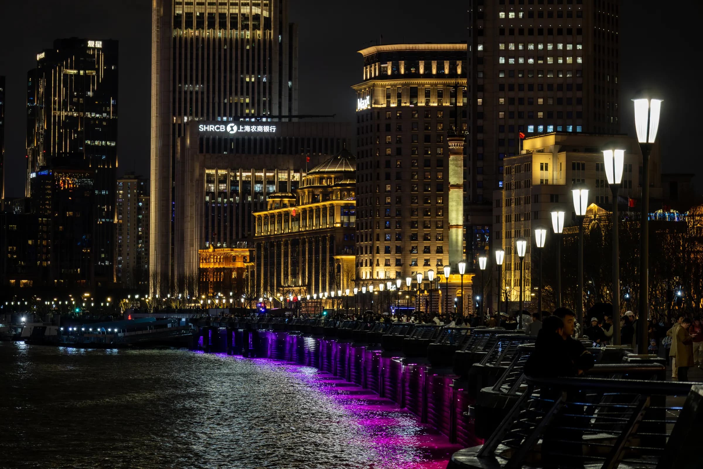 The Bund at night from the river wall: lit heritage buildings, a row of street lamps and violet light along the water's edge.