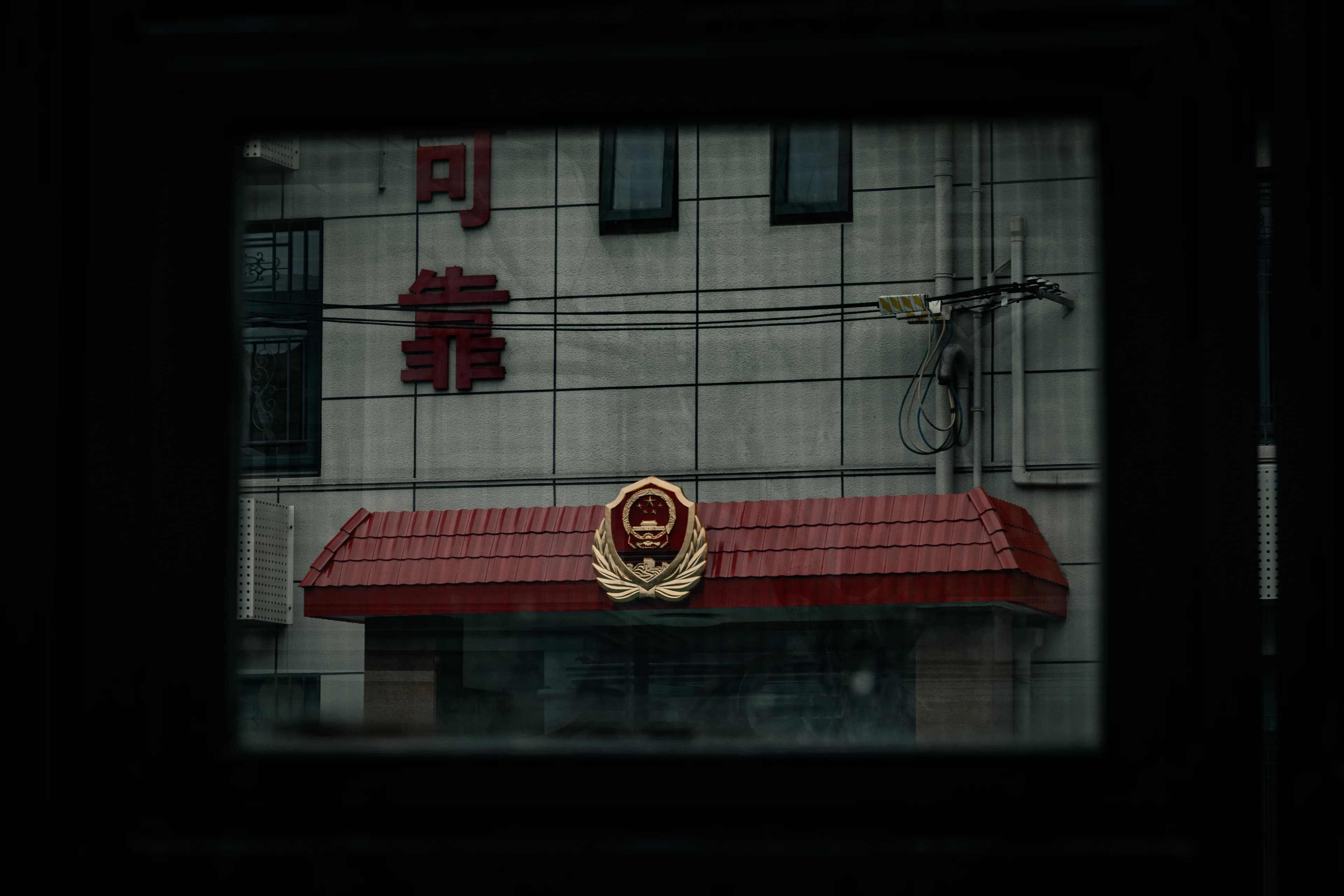 Through a dark window frame, a small red-tiled gateway topped with a gold emblem, red characters on the white tiled wall above it.