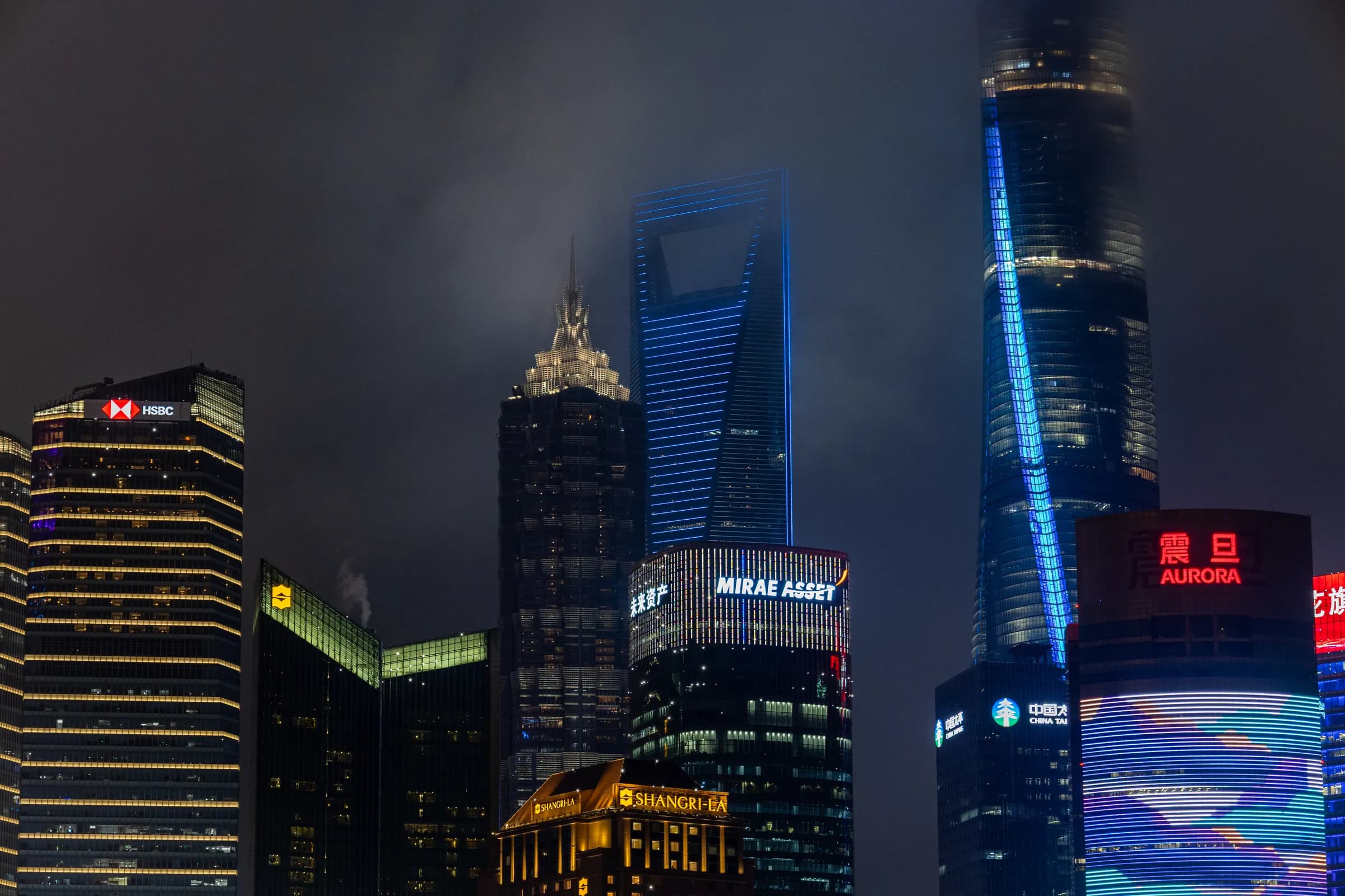 Lujiazui's towers at night with their tops lost in low cloud: the Shanghai Tower's blue light strip, the World Financial Center and lit screens below.