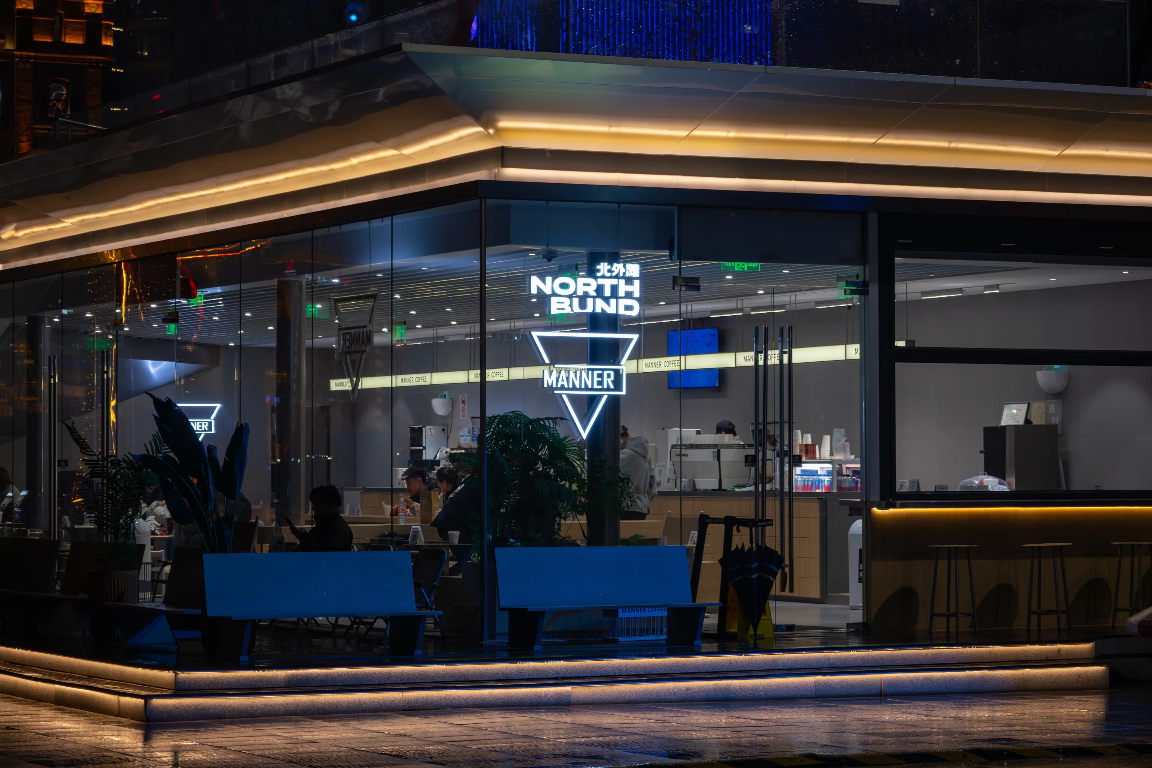 A glass-walled café on the North Bund at night, warm light strips along its roof edge and a glowing triangle logo inside.