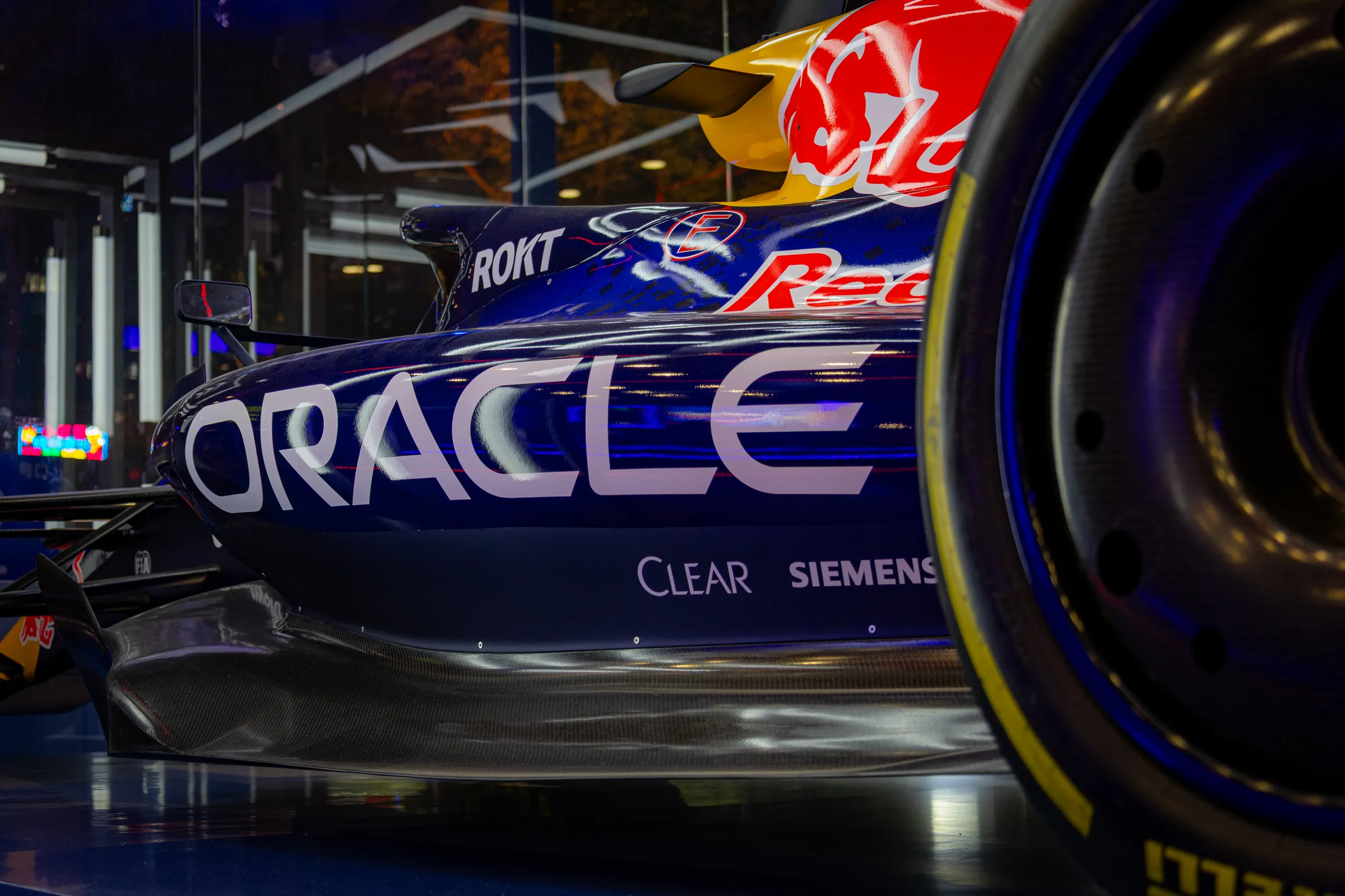 The side of a Red Bull Formula One car in a showroom window, ORACLE in large white letters along the sidepod beside a rear tyre, street lights reflected in the glass.