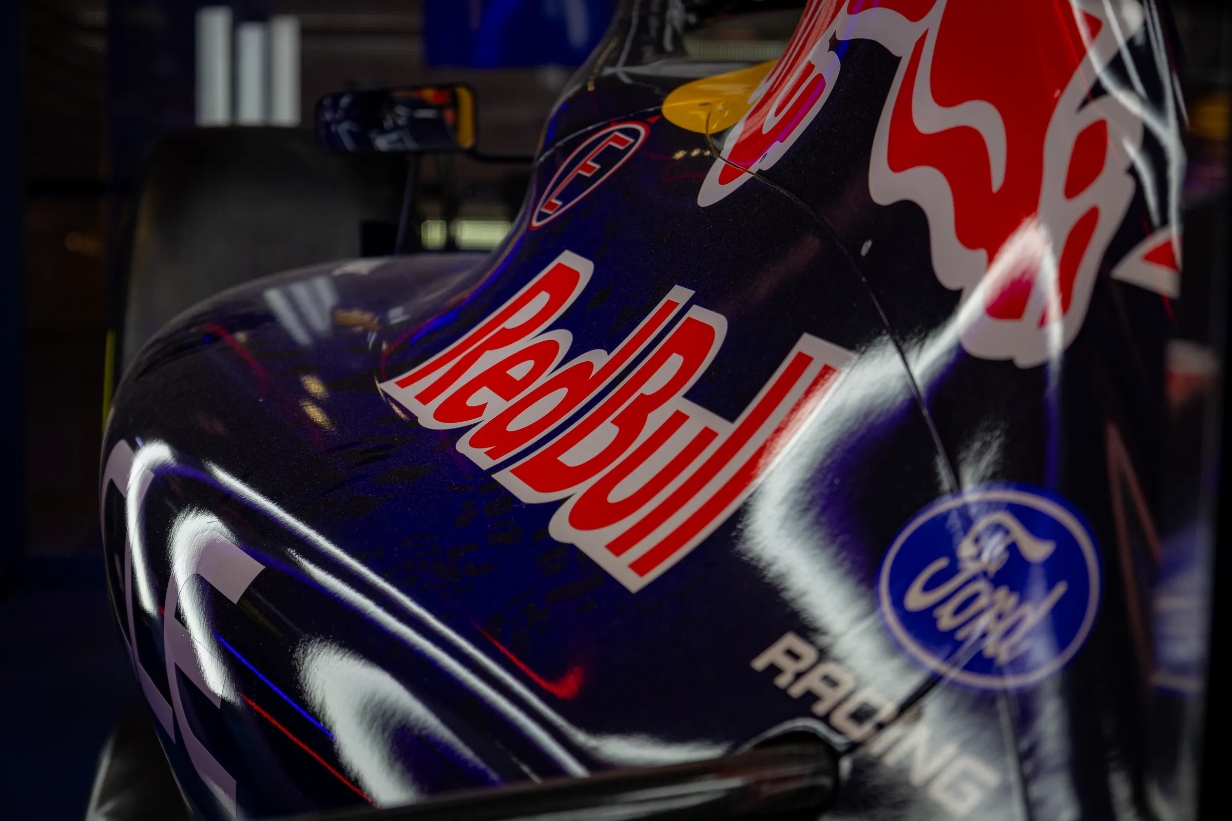 Close-up of a Red Bull Formula One car's engine cover in a dim showroom, the red Red Bull lettering and a Ford Racing badge catching the light on the dark blue bodywork.