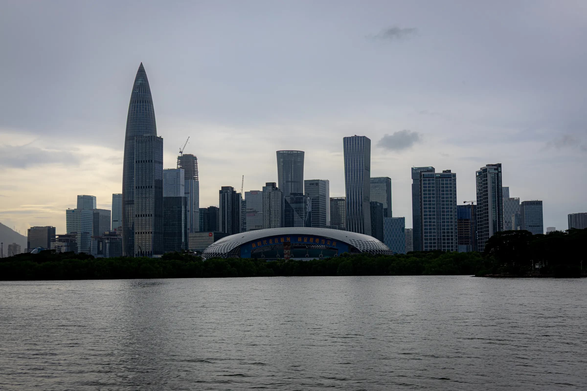 Shenzhen towers across still grey water on an overcast afternoon, the tallest tapering to a point.