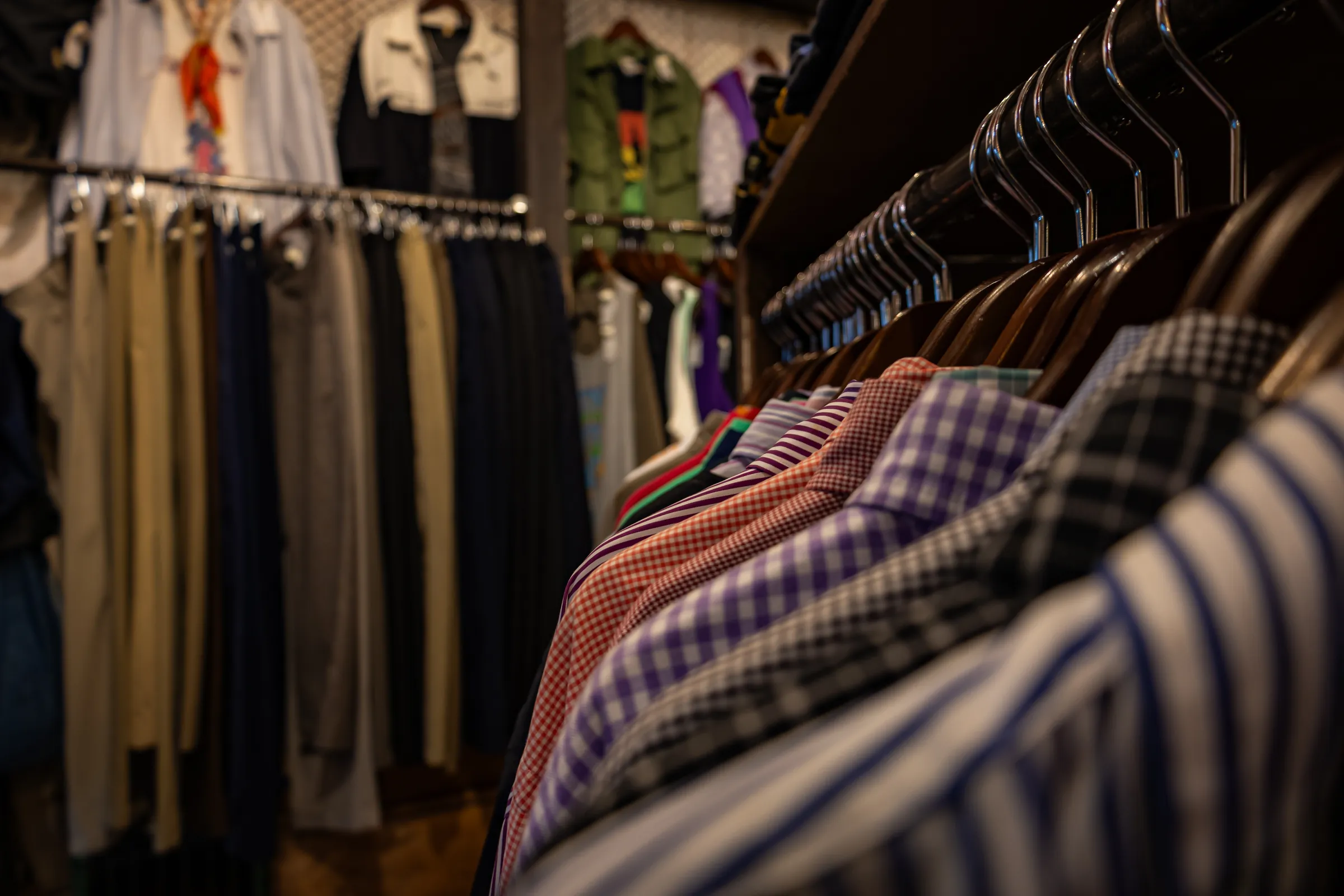 A rack of second-hand shirts in a dim vintage shop, striped and checked collars lined up in the foreground and more clothes hanging on rails behind.
