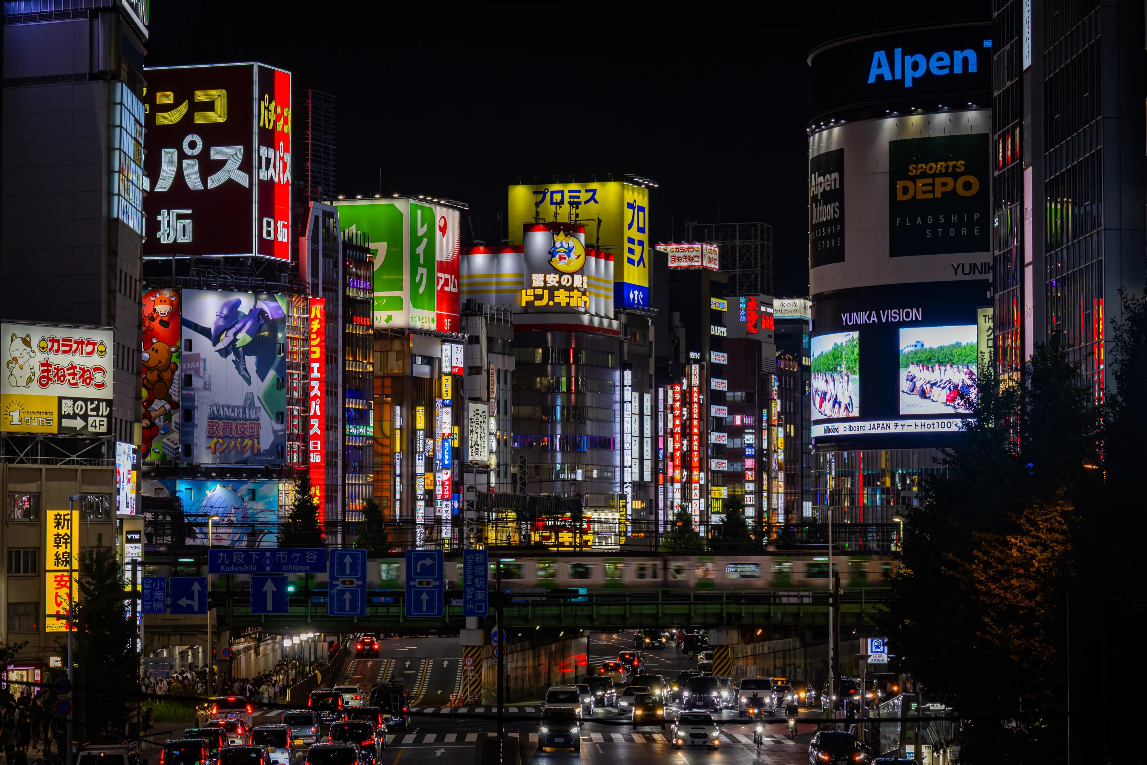 Shinjuku at night: stacked neon and video signs above a street of traffic and an elevated railway.