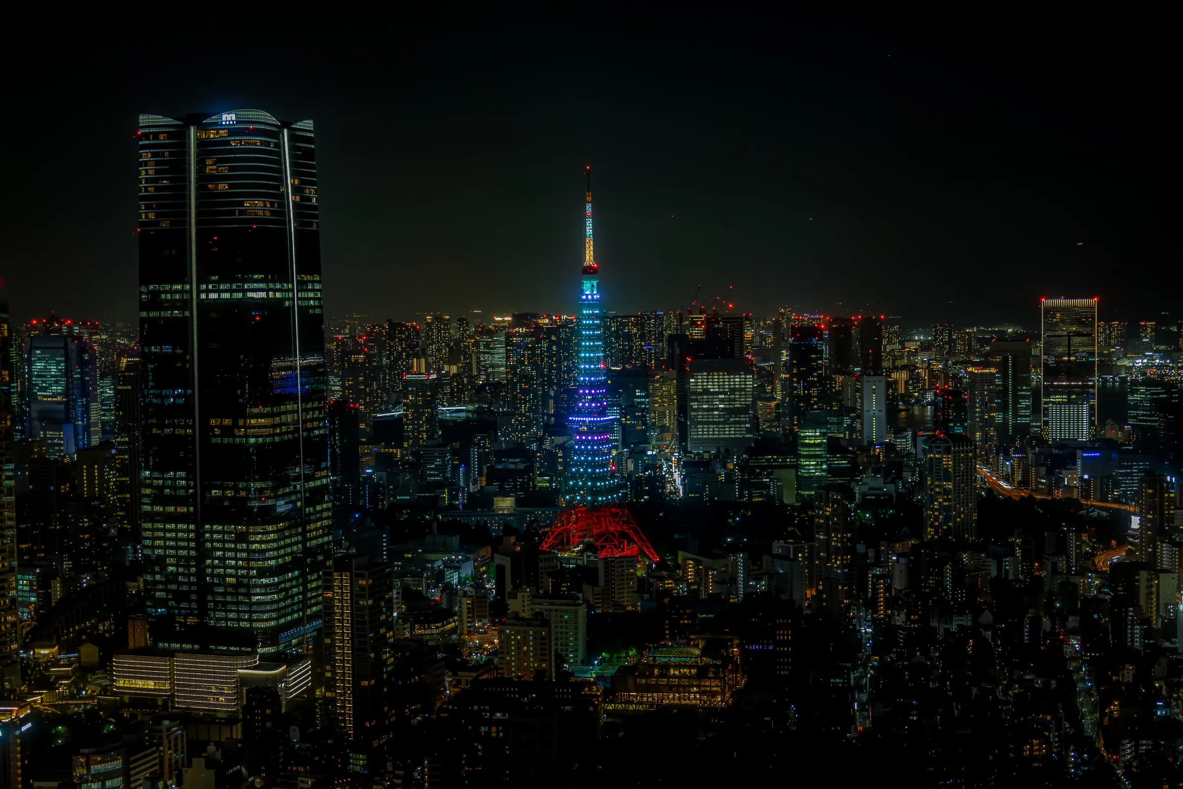 Tokyo from high above at night: Tokyo Tower lit blue in the middle of the city, a dark glass skyscraper on the left.