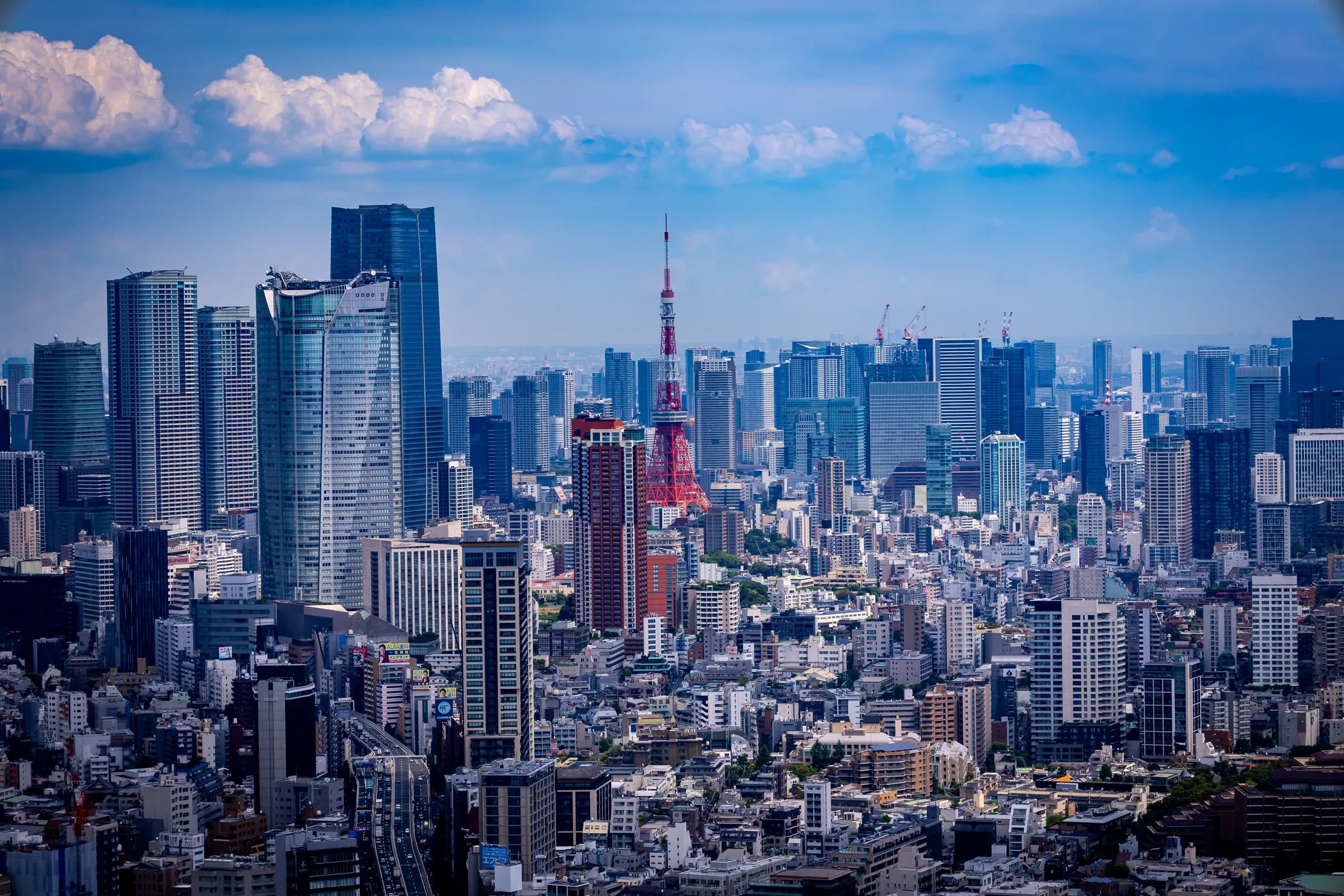 Tokyo by day from above, Tokyo Tower small and red among glass skyscrapers under summer clouds.