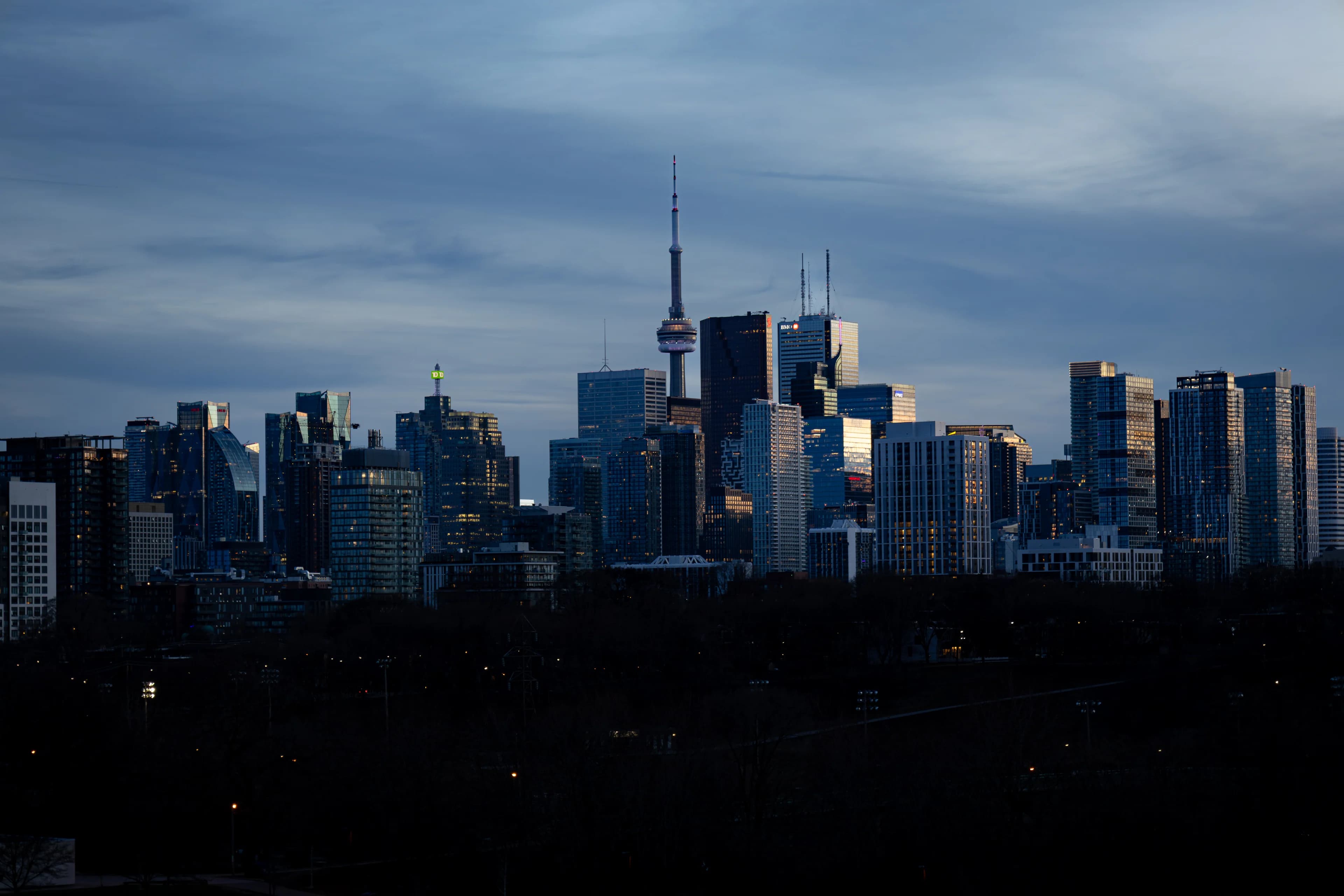 Downtown Toronto at dusk under heavy slate-blue cloud, the CN Tower rising from the middle of the towers above a dark band of trees.