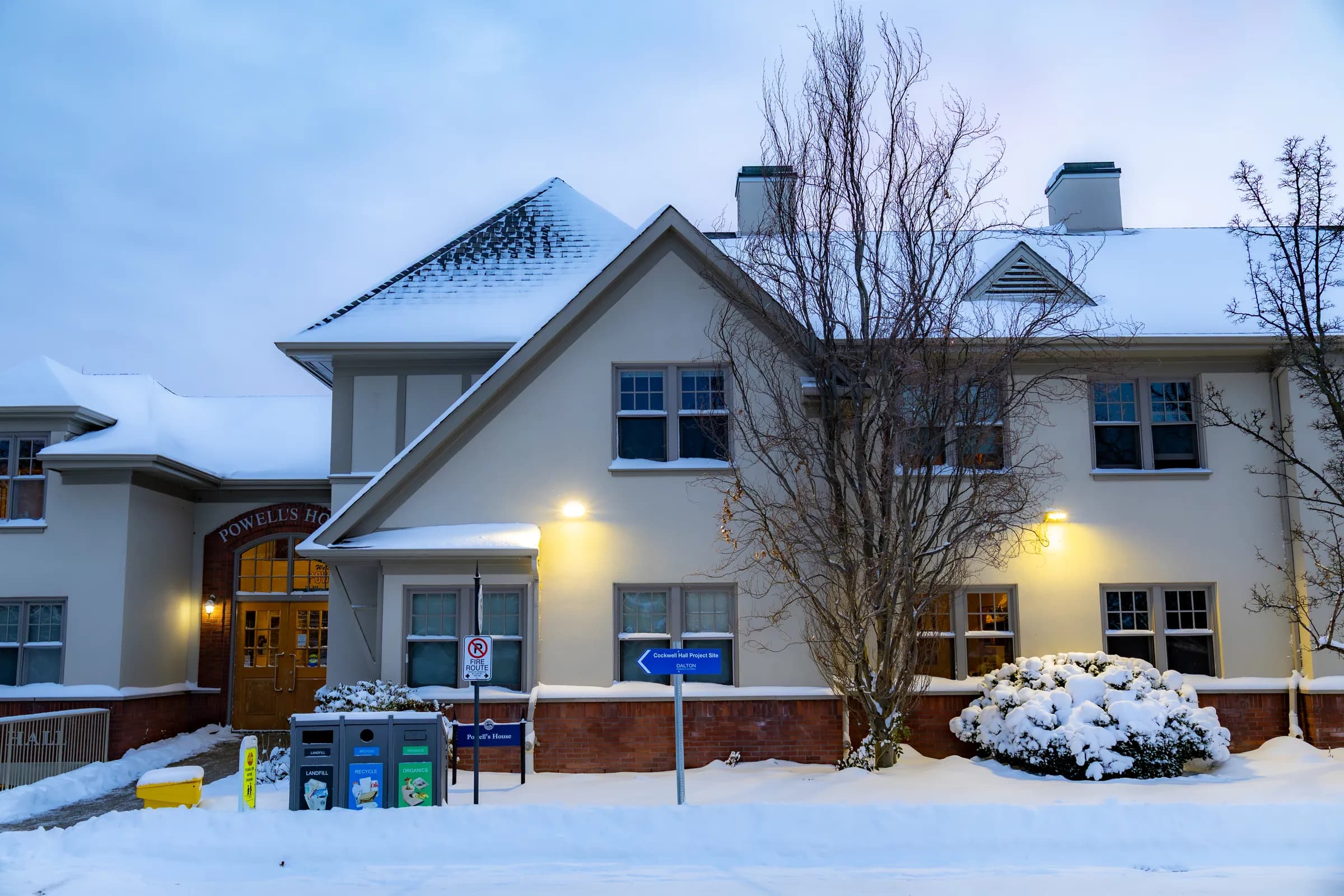 A white house with warm porch lights after a fresh snowfall, snow piled on the roof, the shrubs and the lawn.
