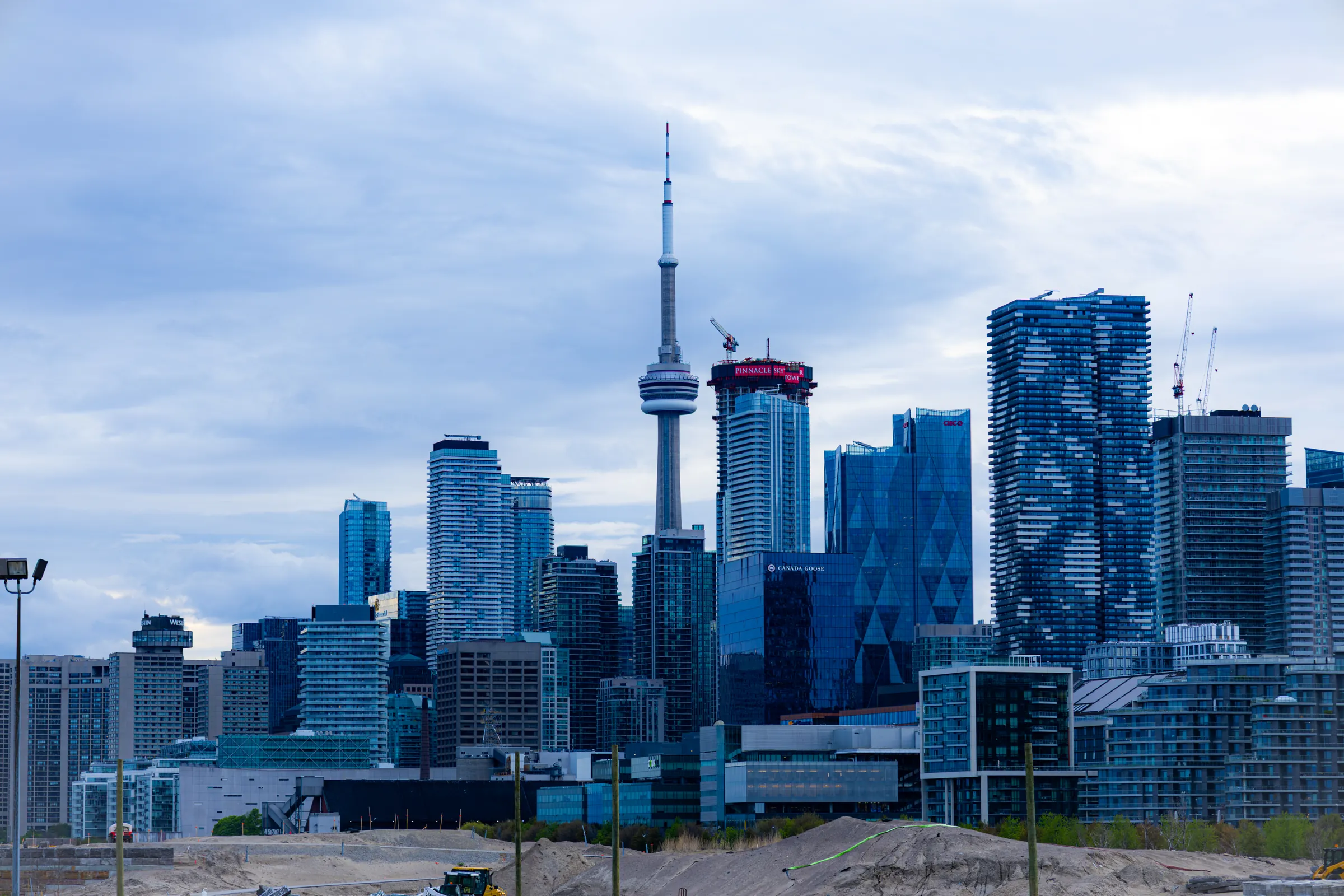 Toronto's skyline on an overcast evening, the CN Tower among glass condo towers, low waterfront buildings and a stretch of construction sand in front.