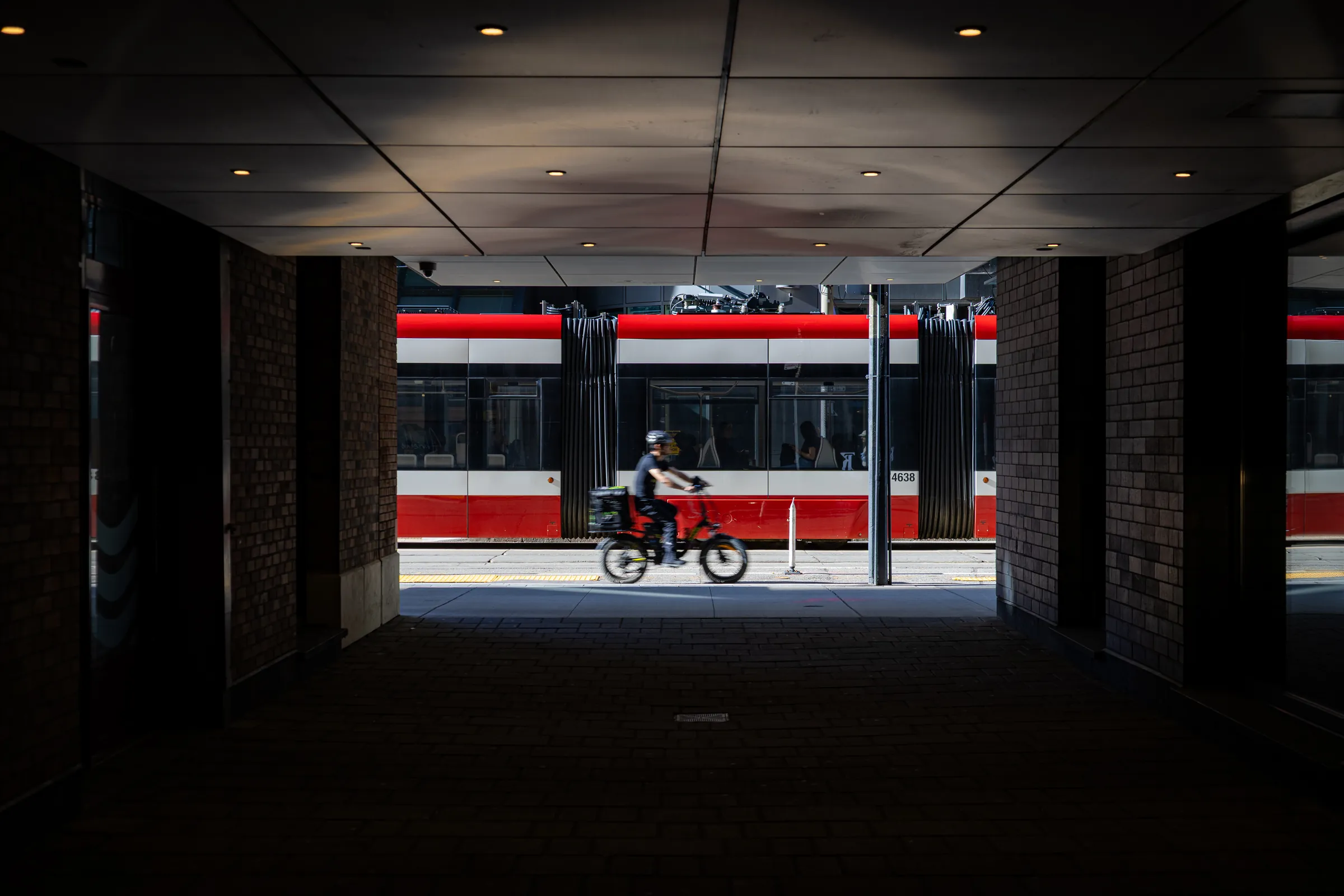 From inside a dark covered passage, a red-and-white Toronto streetcar passes across the bright opening, a cyclist riding in front of it.