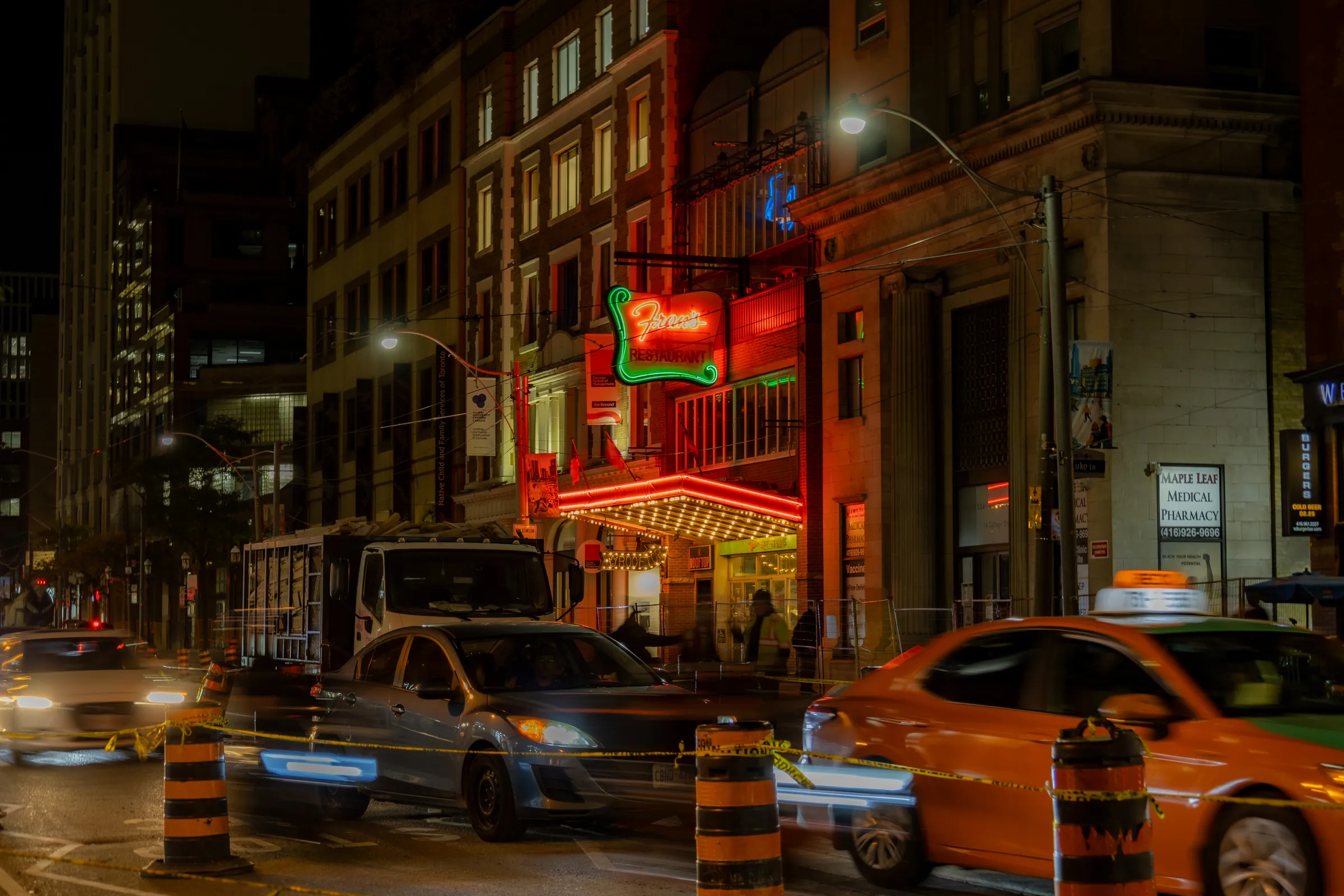 A downtown Toronto street at night, an orange taxi and other cars slightly blurred past construction barrels, a brick building with red neon signs behind them.