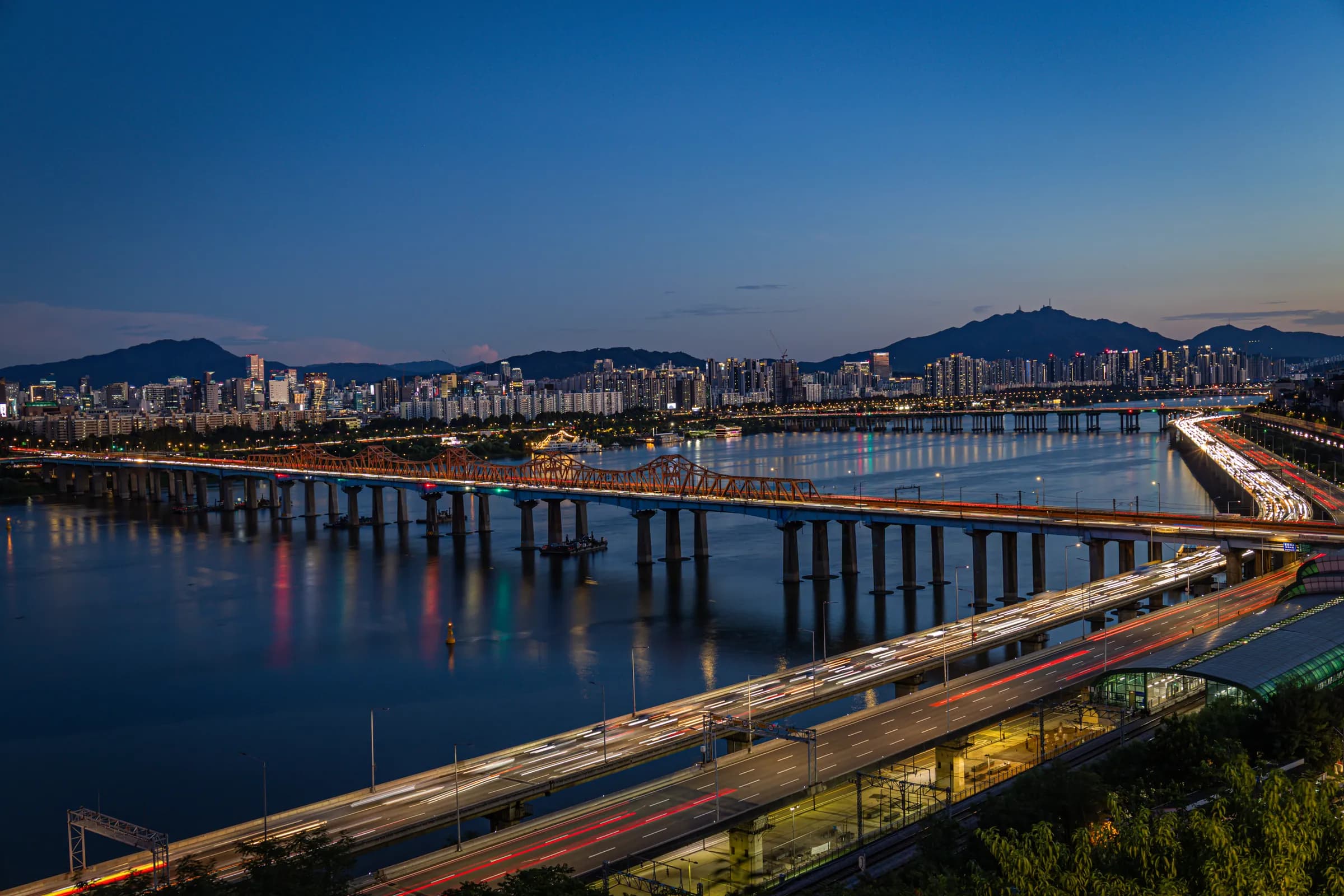 Blue-hour view over the Han River in Seoul, with a long lit bridge, light trails on a riverside expressway and mountains on the horizon.