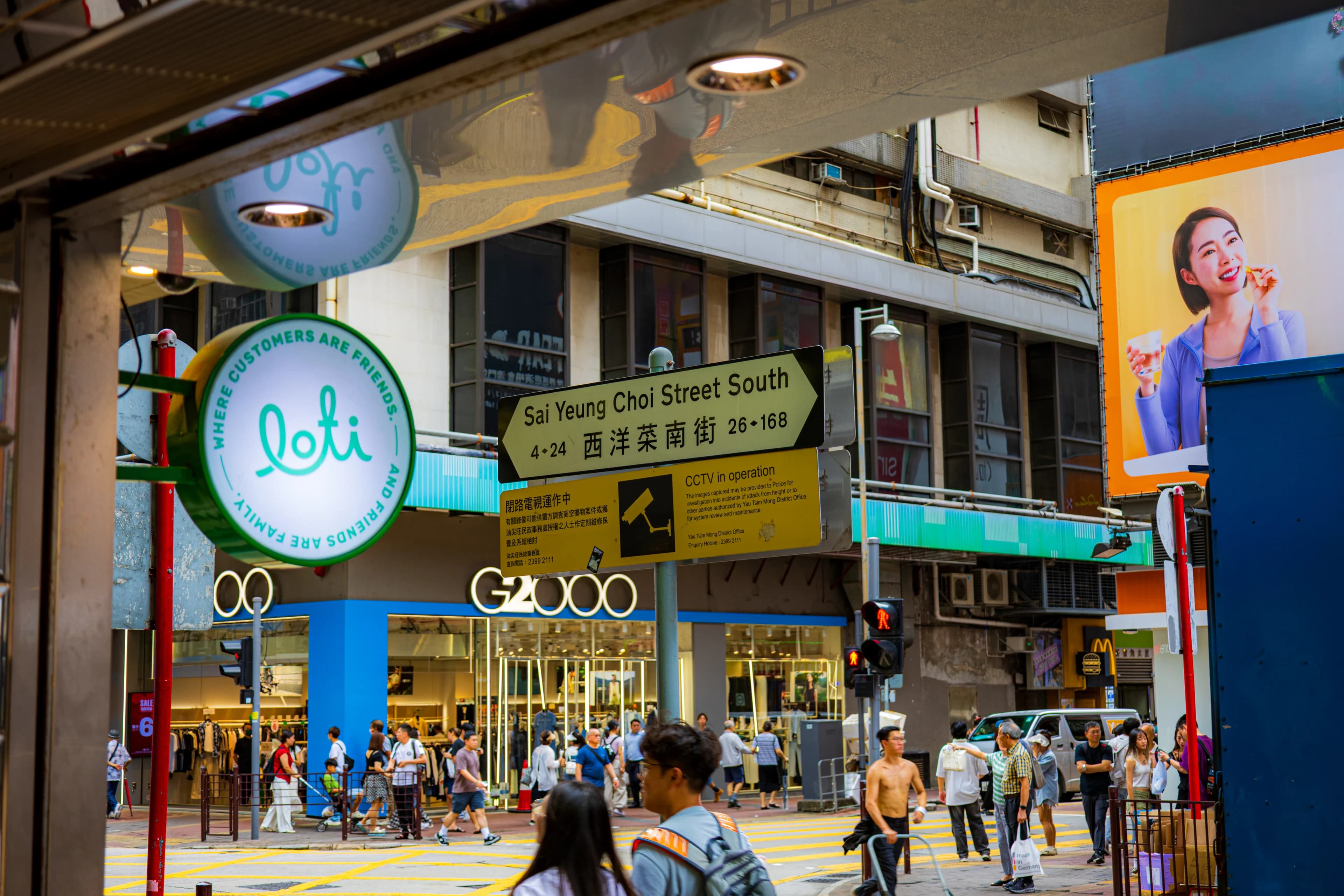 A busy crossing on Sai Yeung Choi Street South in Mong Kok by day: a yellow street sign and CCTV notice in the middle, shoppers crossing below in front of a G2000 store, a round café sign at left and a billboard at right.
