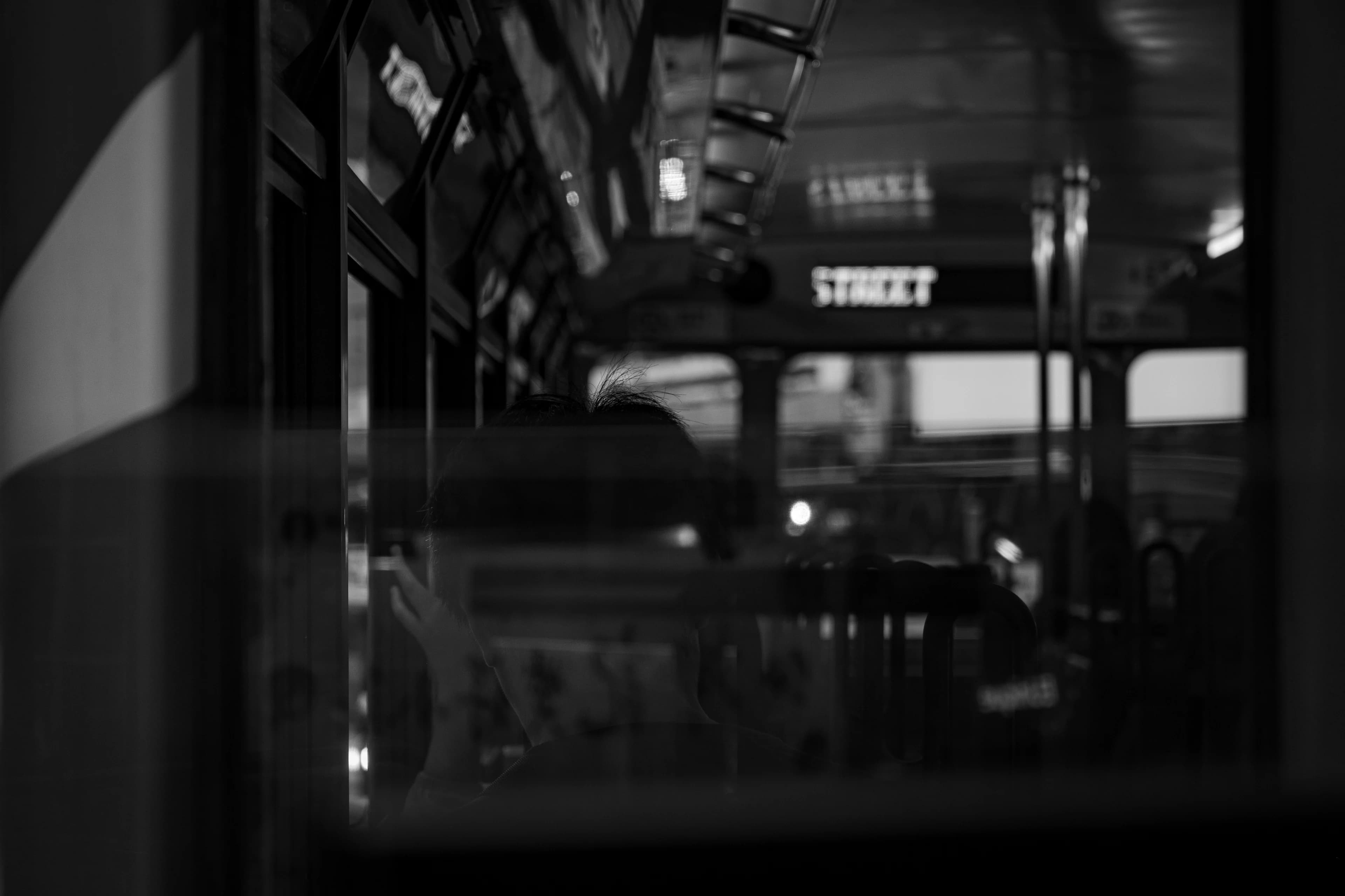A black-and-white view through a Hong Kong tram window at night: a passenger in silhouette in front, the lit STREET destination board sharp beyond the blurred rails and windows.