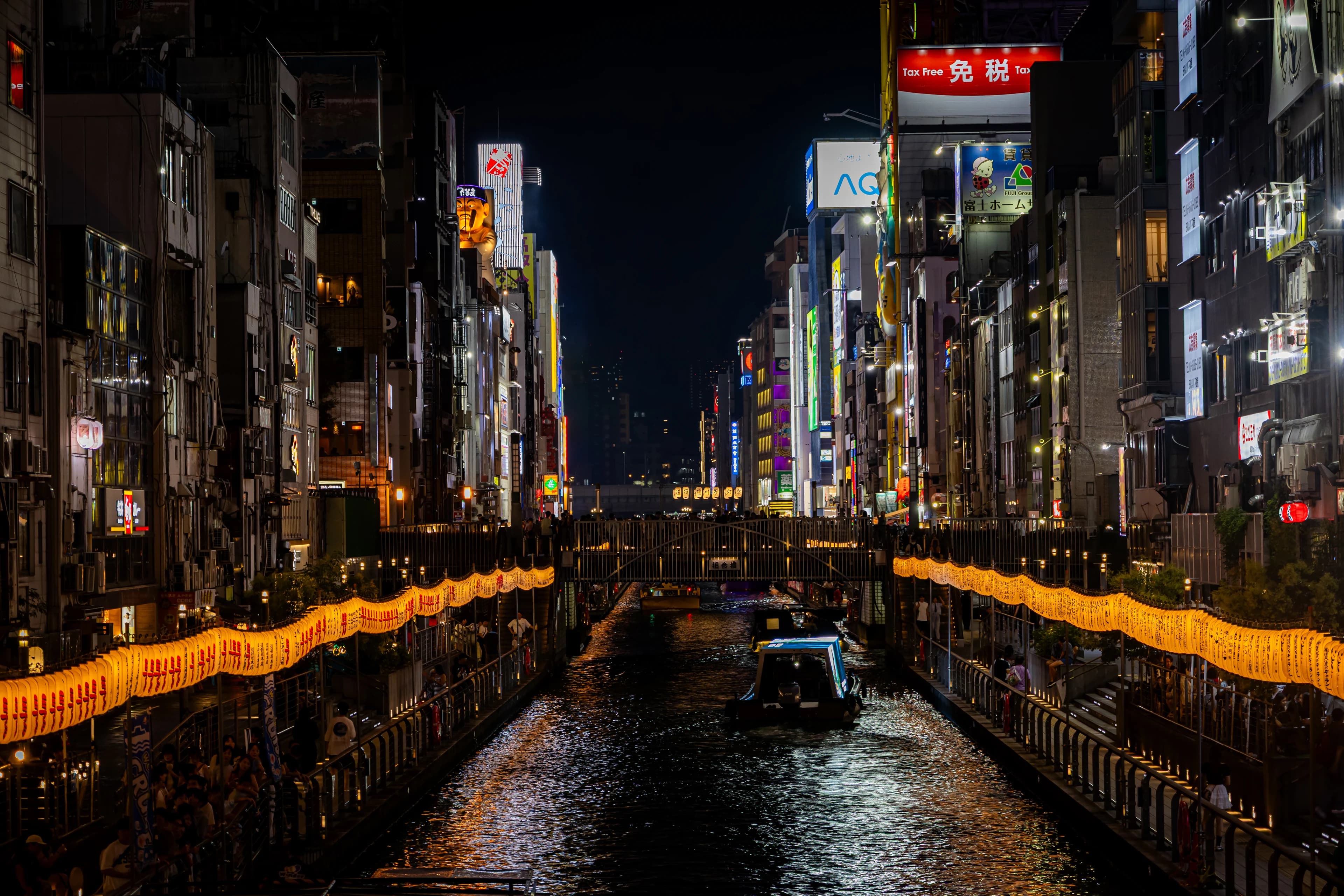 The Dotonbori canal at night, lanterns glowing along both banks under walls of neon, a tour boat in the water.
