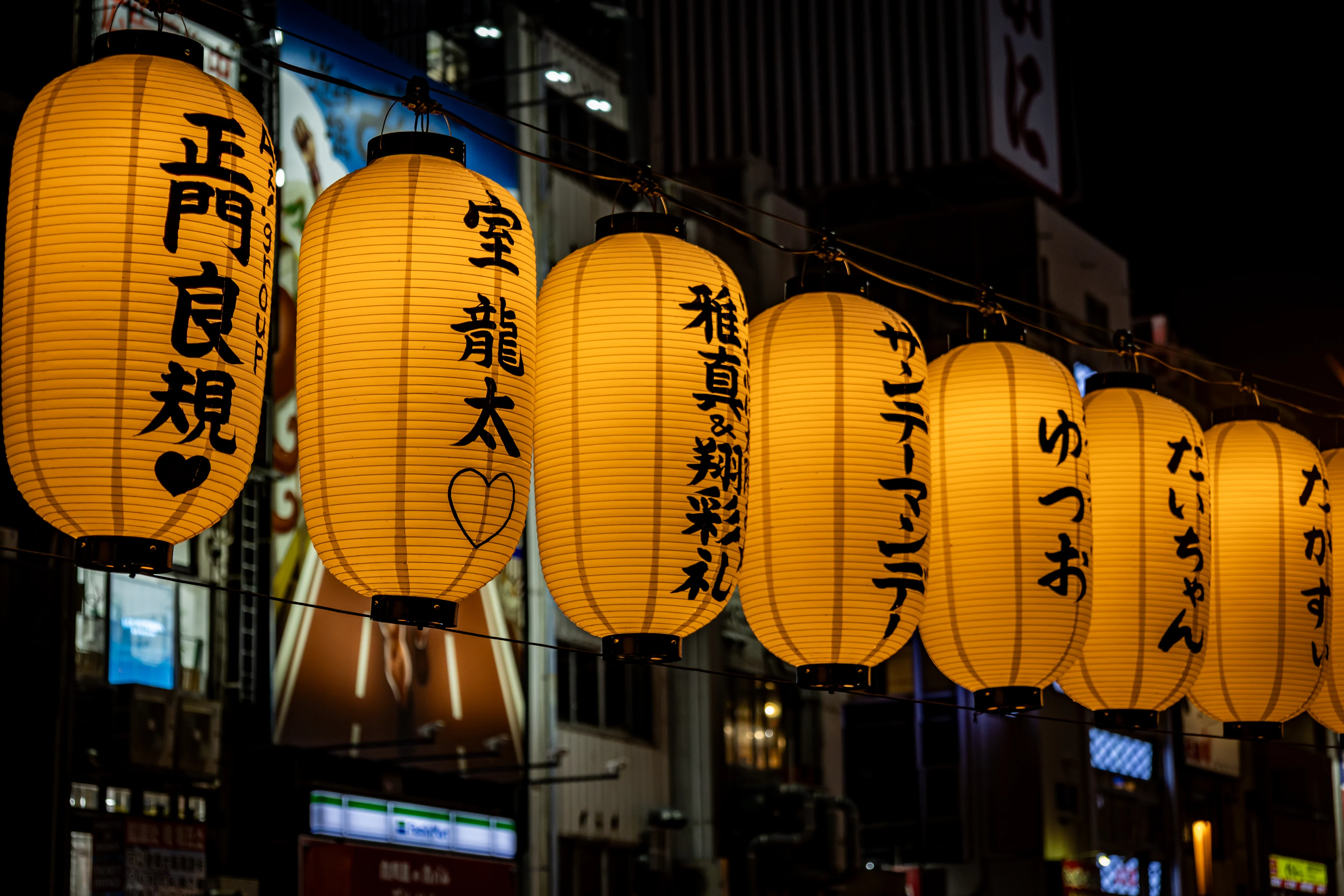 A row of glowing yellow paper lanterns in Dotonbori, each painted with names in black brush strokes.