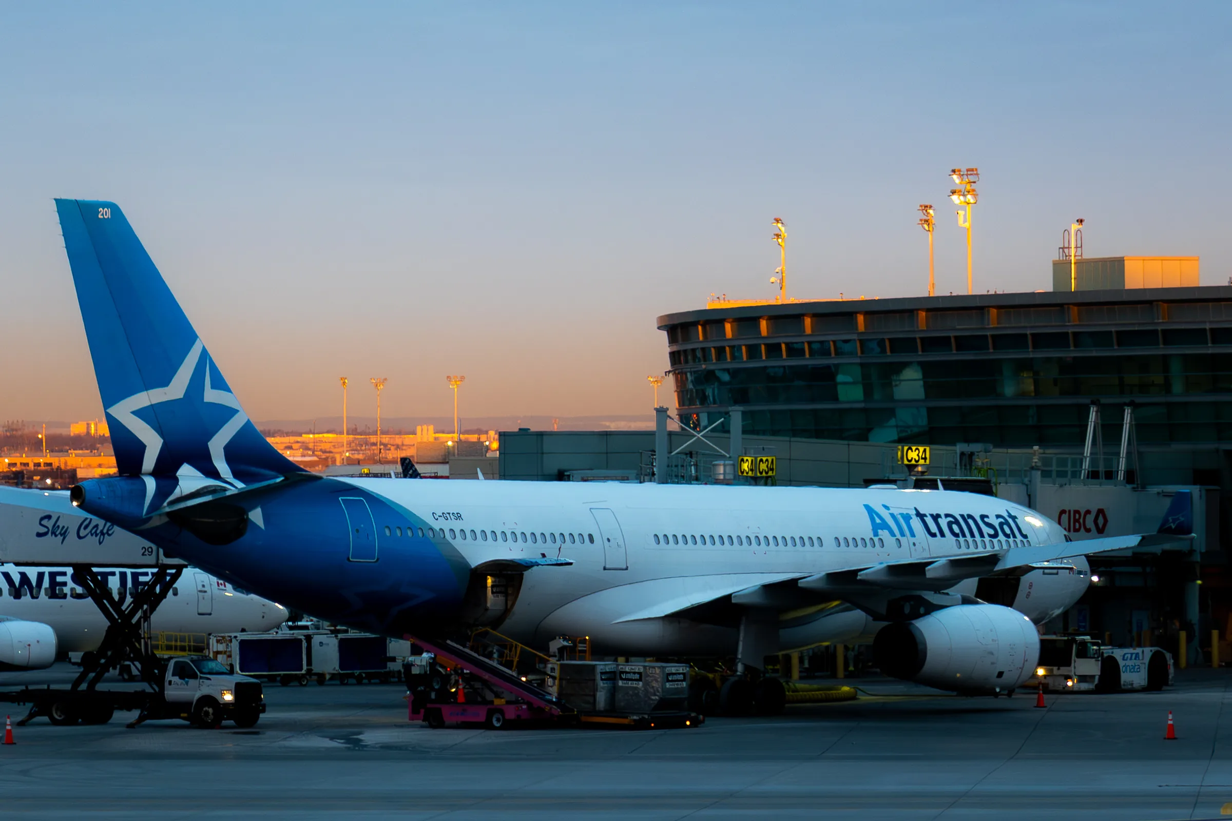 An Air Transat widebody parked at a Pearson gate in early light, its blue star tail against a peach sky, the terminal's glass curve and floodlight masts behind it.
