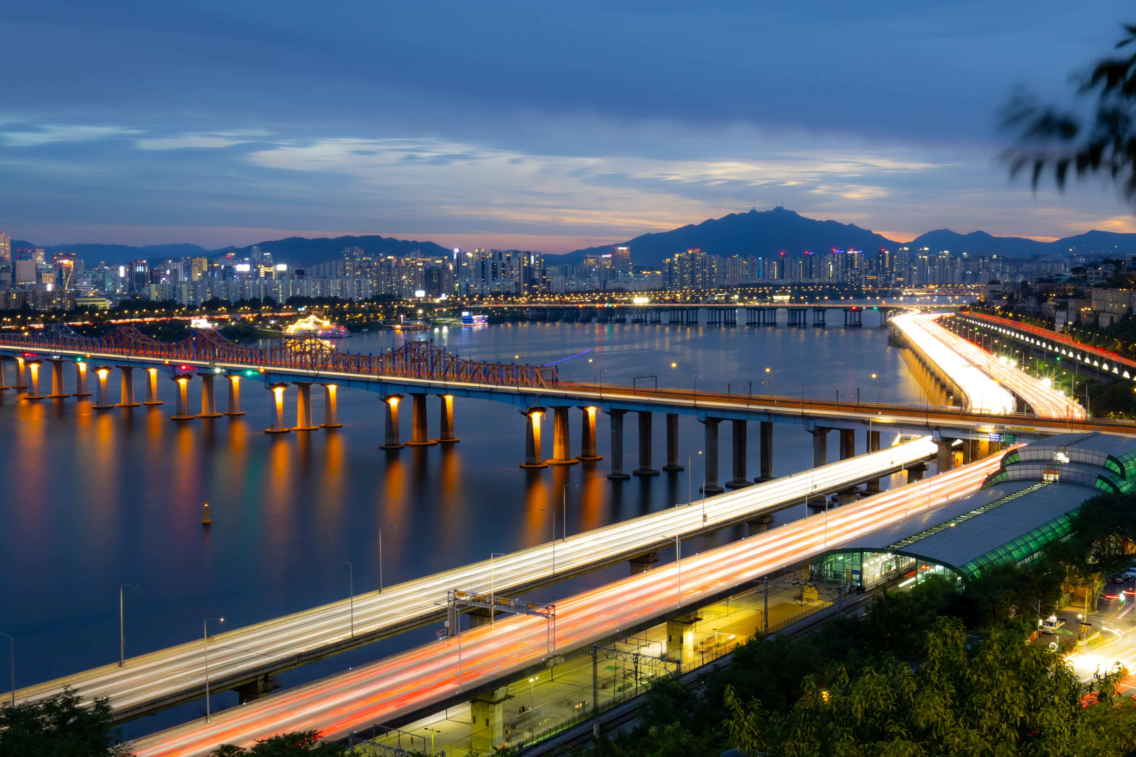The Han at dusk from a wooded hillside: an orange-lit rail bridge crosses the river, a riverside expressway below is drawn out in red and white light trails beside a green-roofed station, and apartment towers and a dark mountain ridge sit under a blue evening sky.
