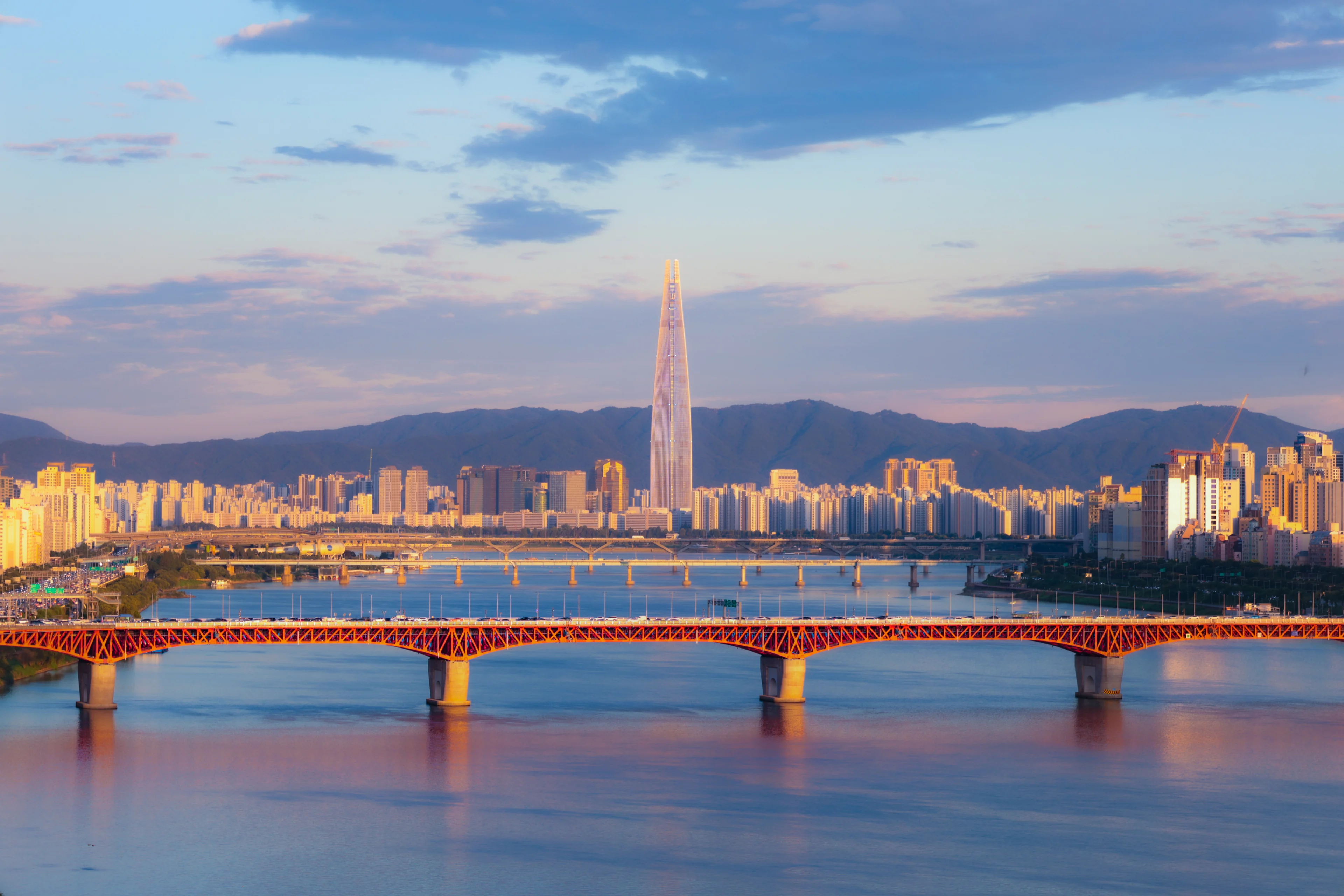 Lotte World Tower catching gold evening light above Jamsil's apartment blocks, mountains behind and a red steel bridge across the Han in front.
