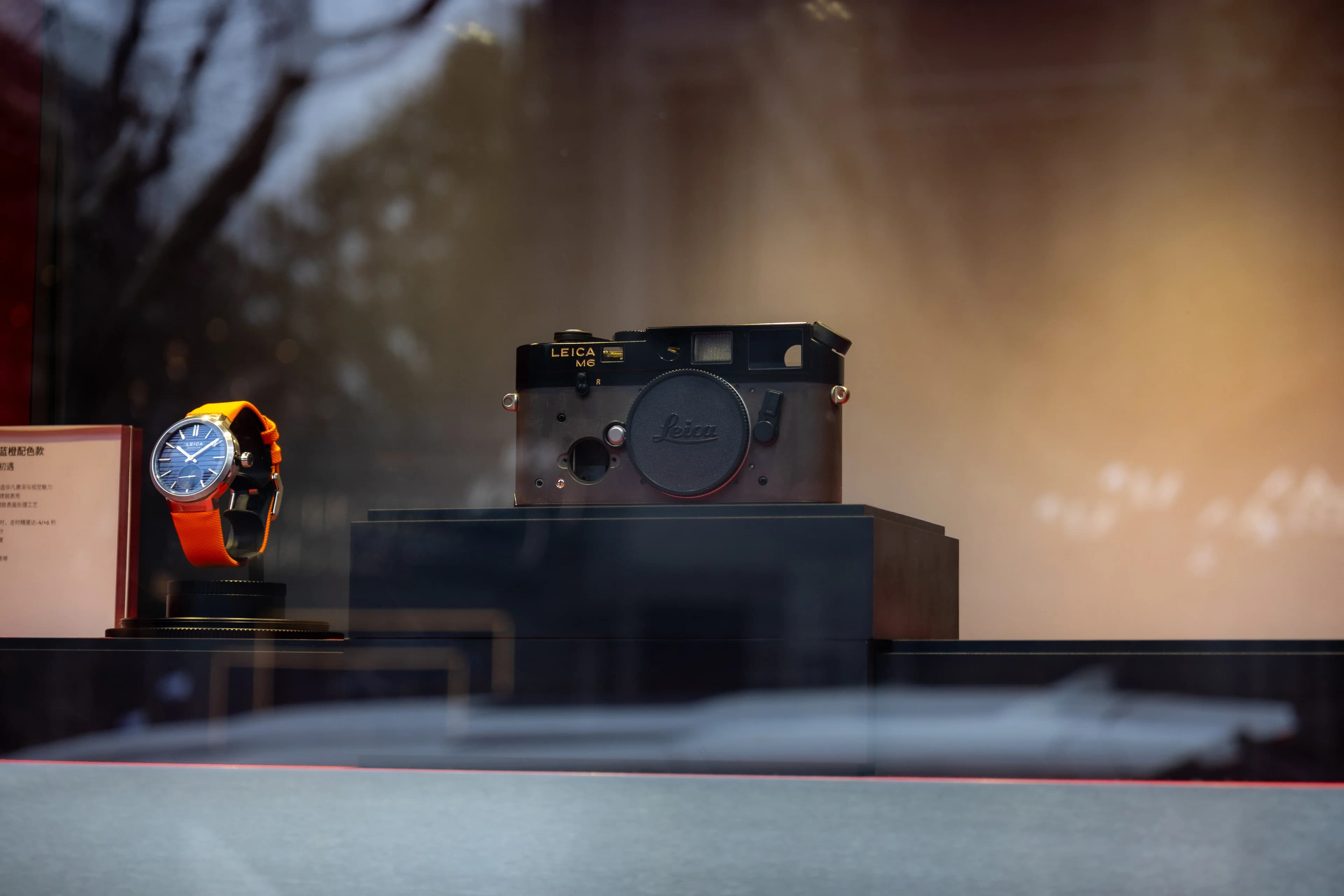 A shop window display: an old Leica rangefinder on a dark plinth beside a watch with an orange strap, the trees and street behind blurred to warm tones.
