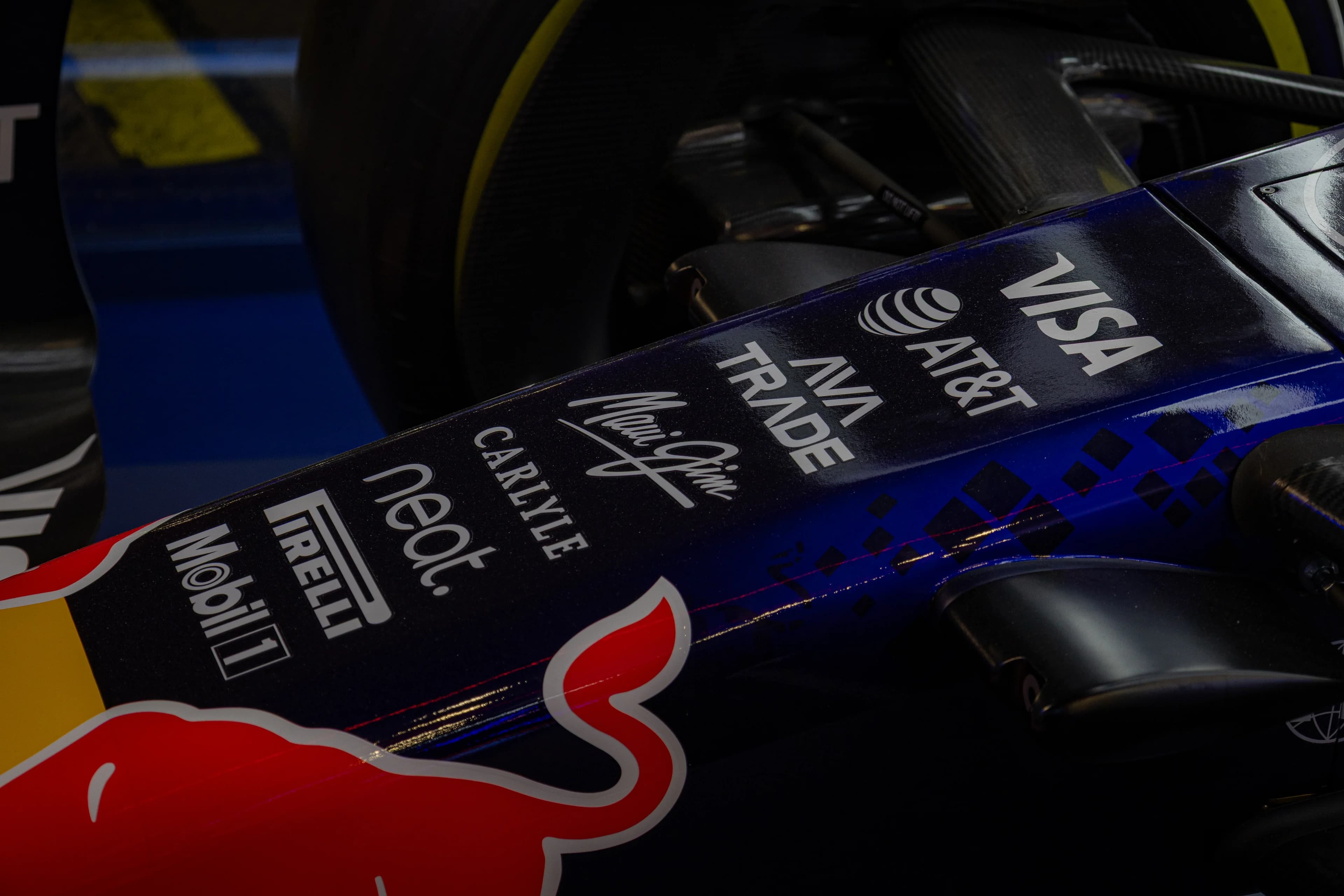 The nose of a Red Bull Formula One car in a dim showroom, its sponsors stacked in white along the dark blue bodywork, Visa and AT&T down to Pirelli and Mobil 1, above the red charging bull.