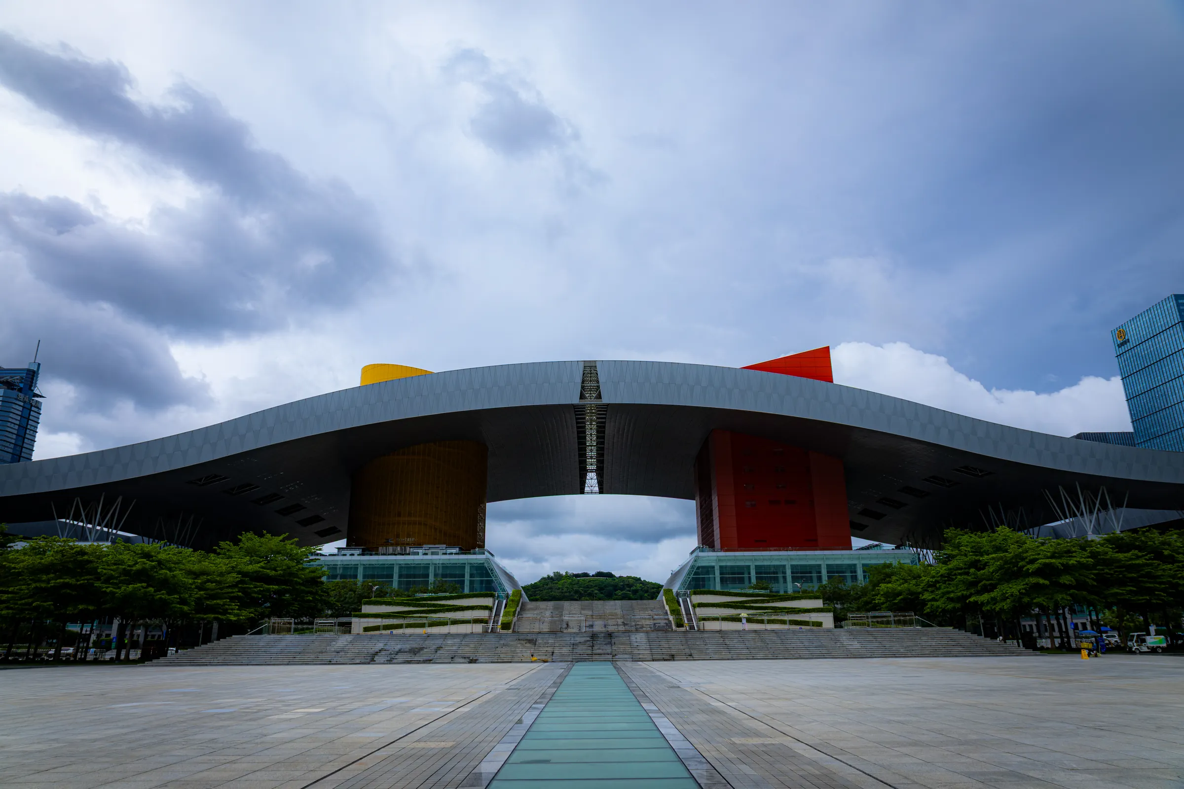 The Shenzhen Civic Center's long winged roof over a wide empty plaza, storm clouds stacked above it.
