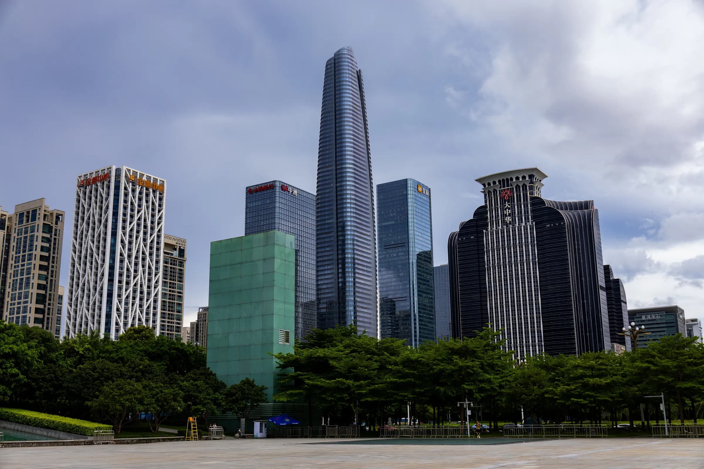 The Ping An Finance Centre rising above Futian's towers on a cloudy day, with a row of trees along a wide plaza.