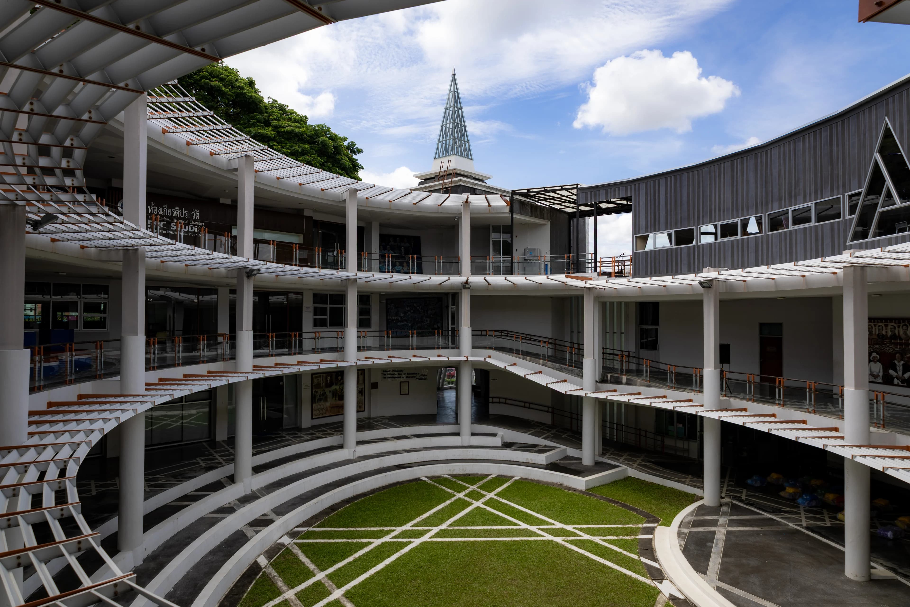 A curved three-storey building of white columns and slatted walkways wrapped around a lawn crossed by white lines, a pointed spire and clouds in a blue sky above.