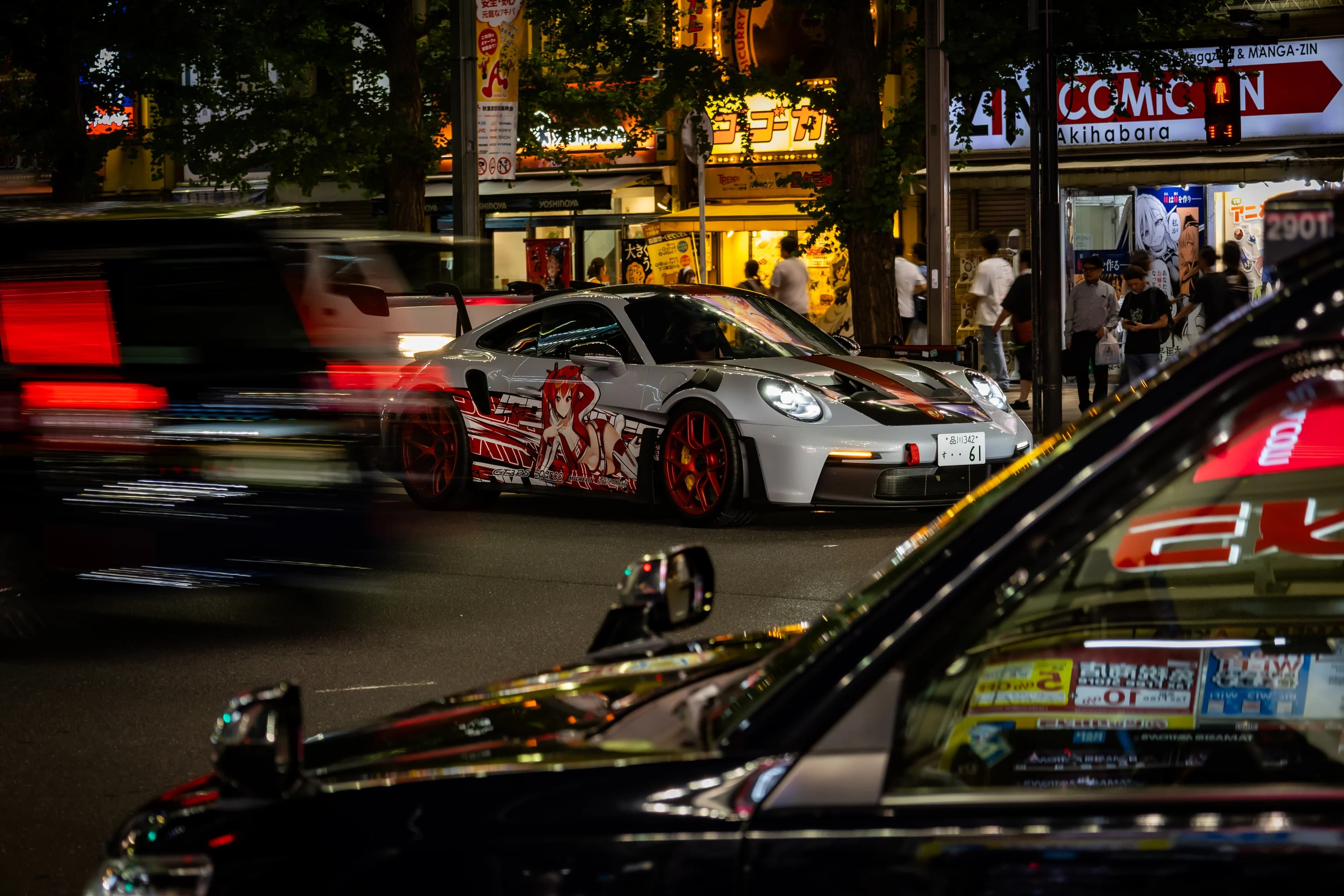 Akihabara at night: a white race-livery Porsche waits at the kerb while traffic blurs past and shoppers crowd under the signs.